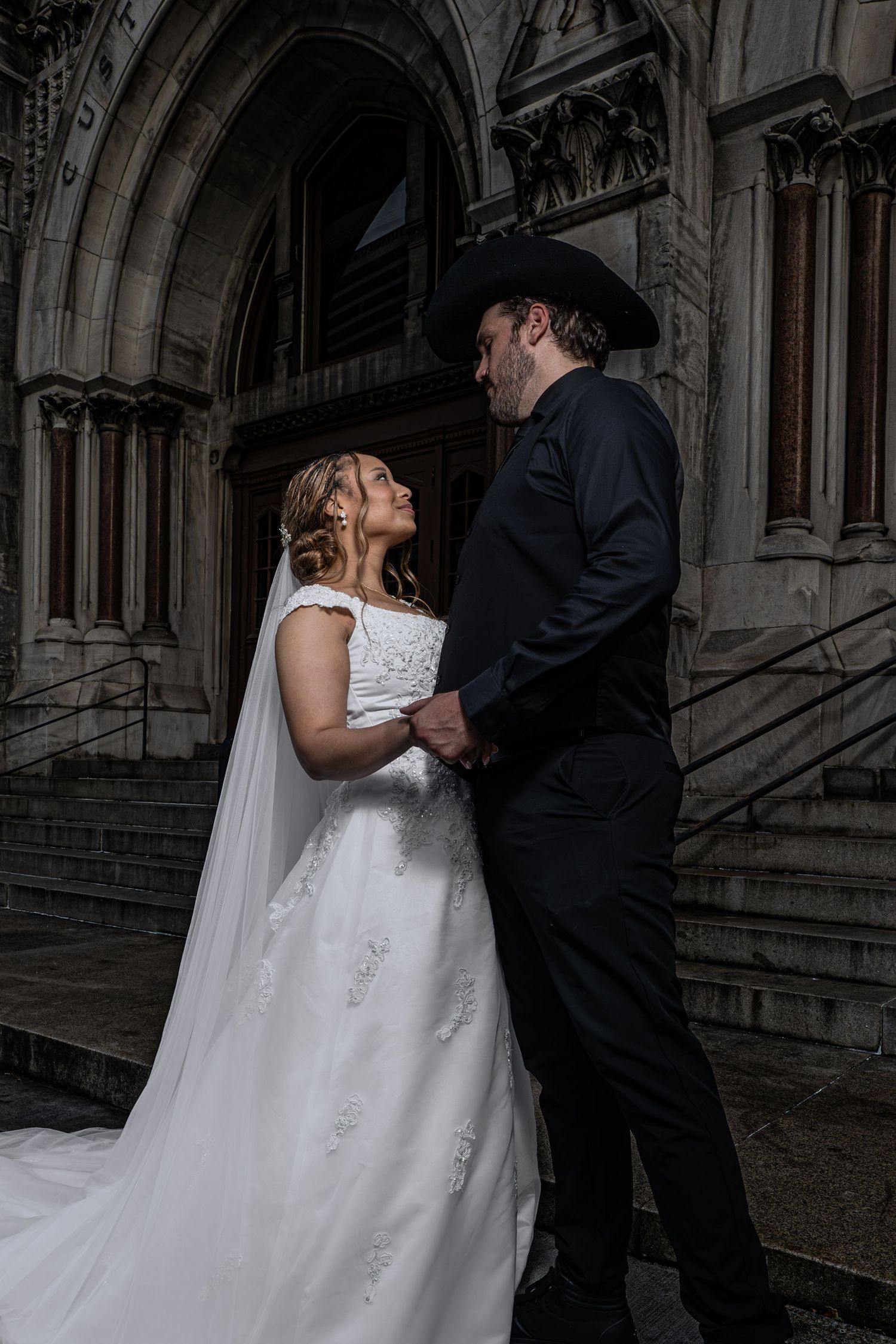 Bride and groom holding hands outside a gothic stone church, groom wearing black suit and cowboy hat.