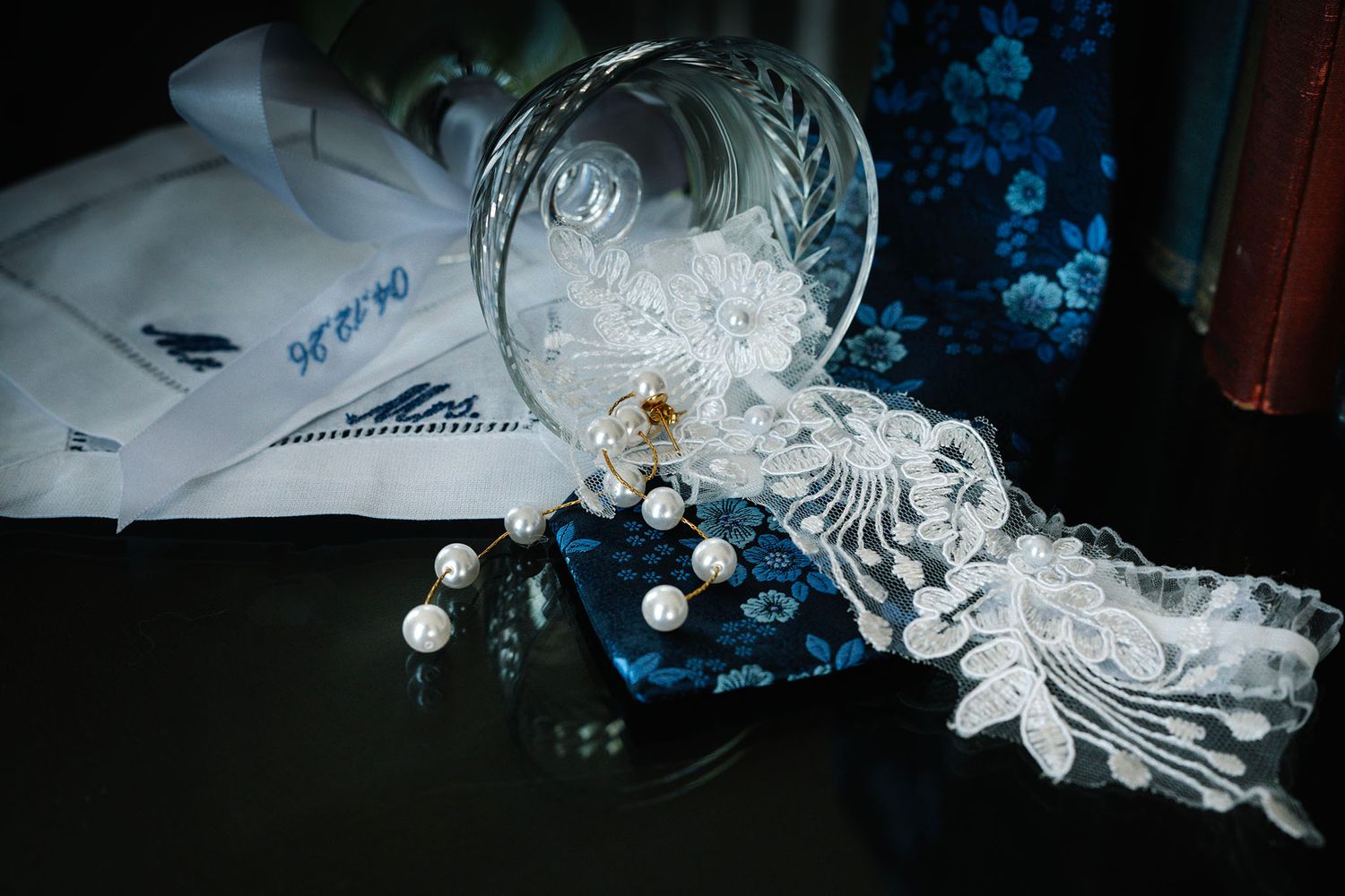 Crystal glass tipped over with pearl jewelry and white lace on dark surface with blue floral fabric in background.