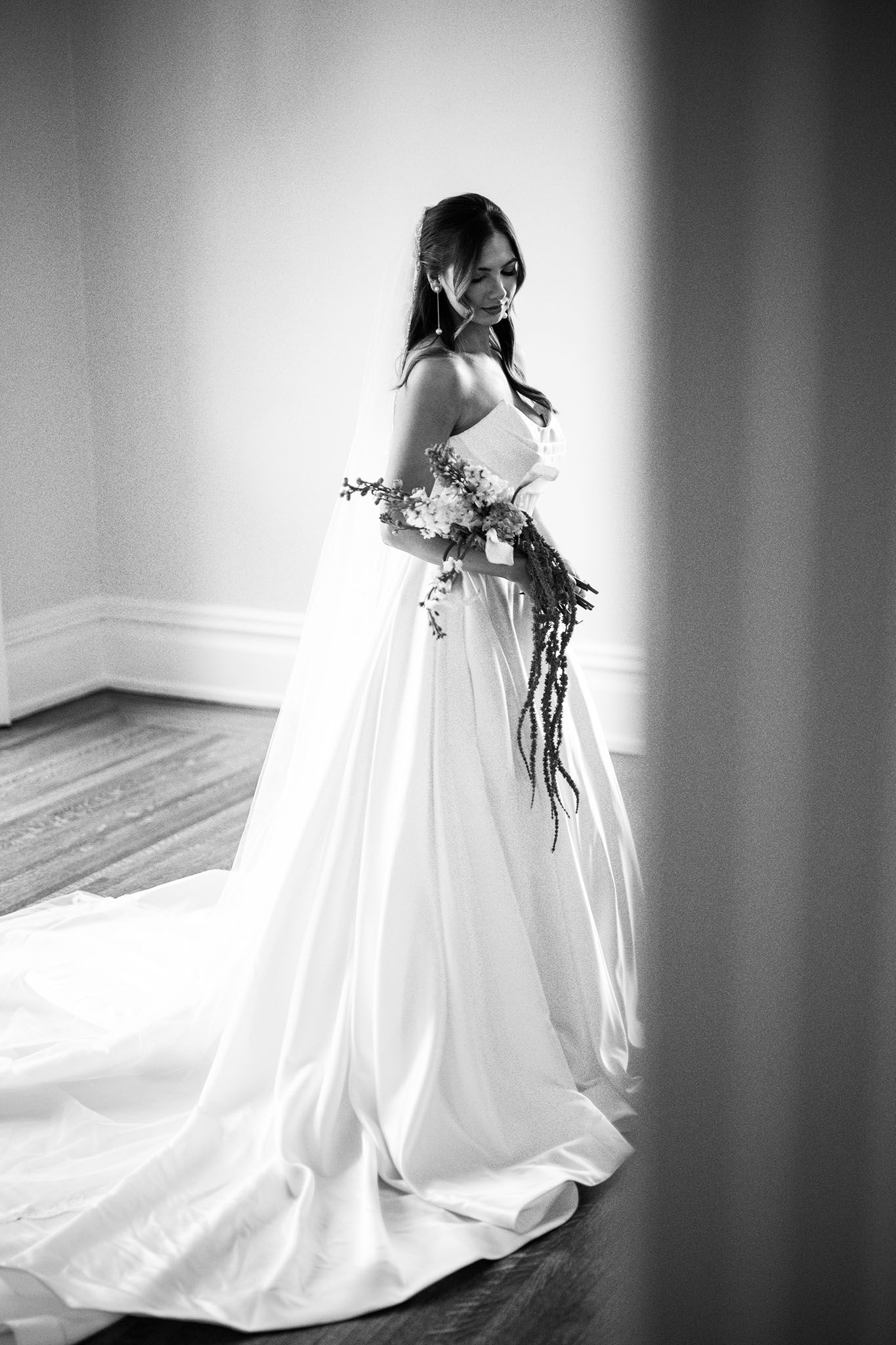Black and white photo of a bride in a flowing strapless gown holding a bouquet, smiling over her shoulder.