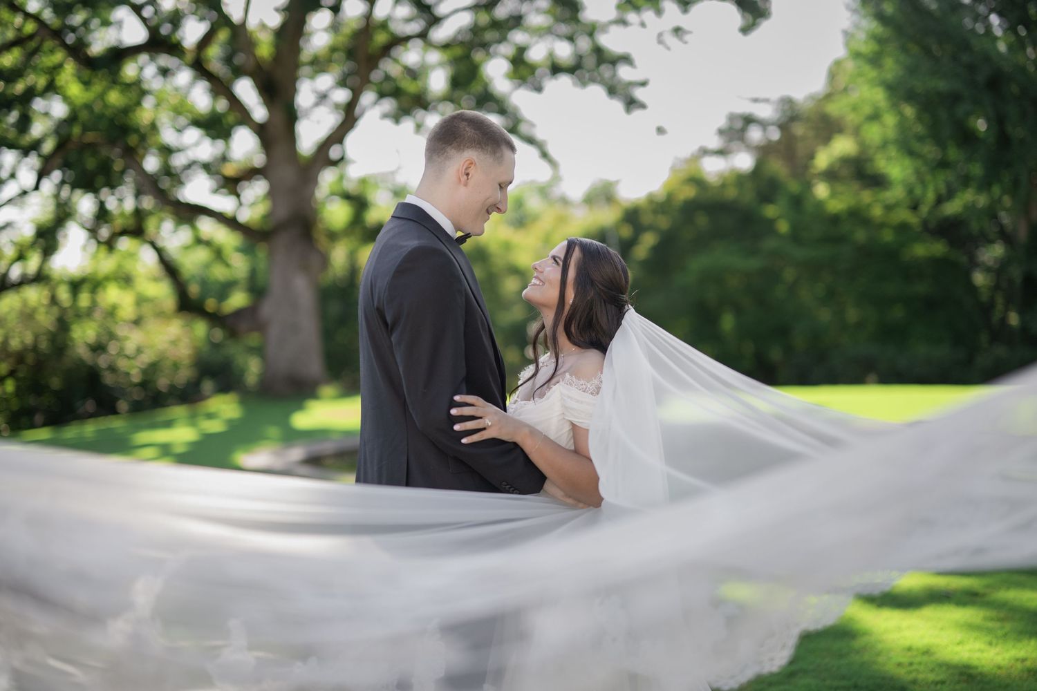 Bride and groom share a romantic moment outdoors as her long veil billows in the wind against lush green trees.
