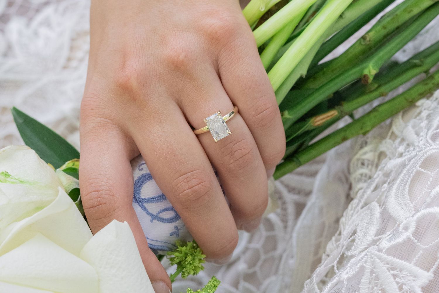 Close-up of a bride's hand wearing a stunning emerald-cut diamond engagement ring while holding a white wedding bouquet.