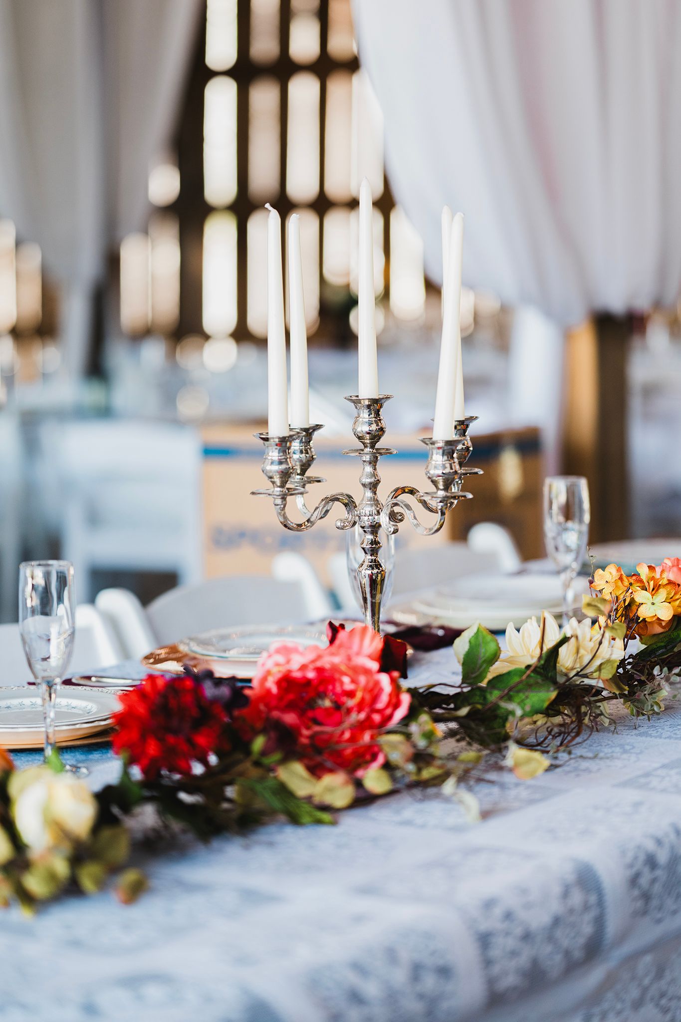 Elegant wedding table with silver candelabra, white candles, and colorful floral garland on a lace tablecloth.