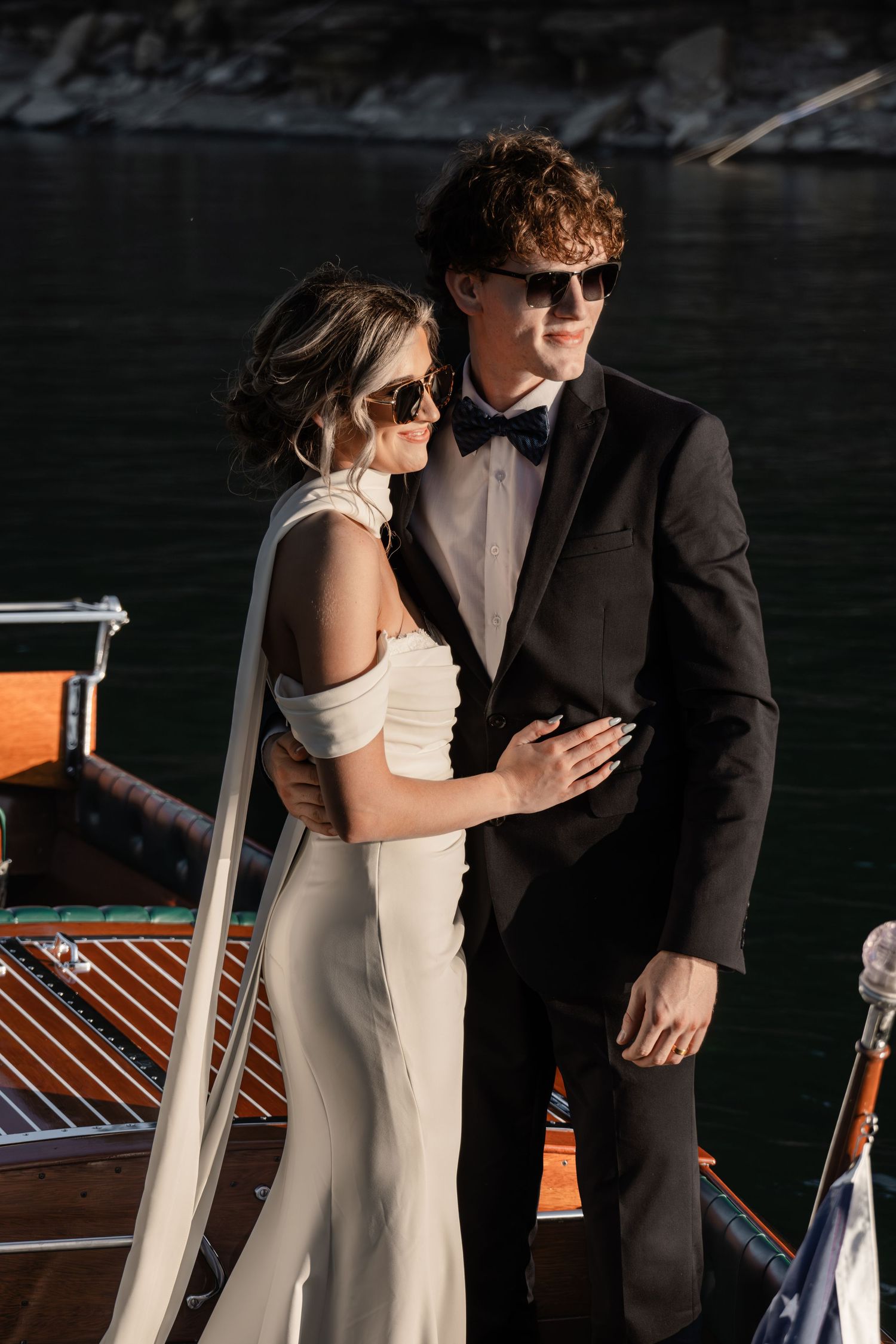 Elegant couple in wedding attire posing on a luxury wooden boat, bride in white gown and groom in black tuxedo with bow tie.