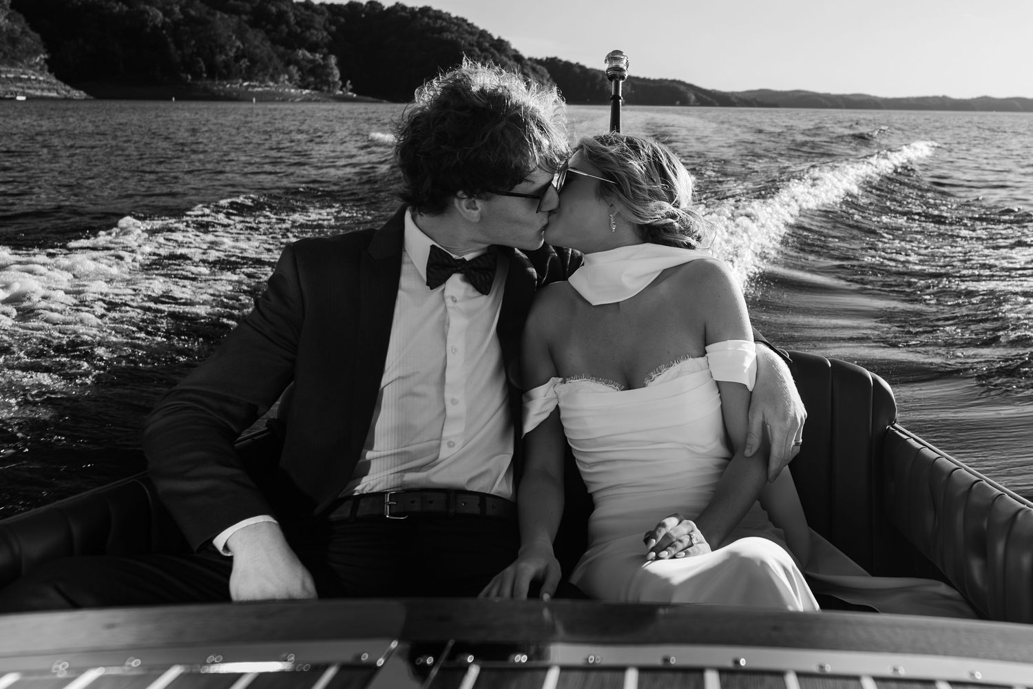 Bride and groom sharing a romantic kiss on a classic wooden boat on the water in a black and white wedding photo.