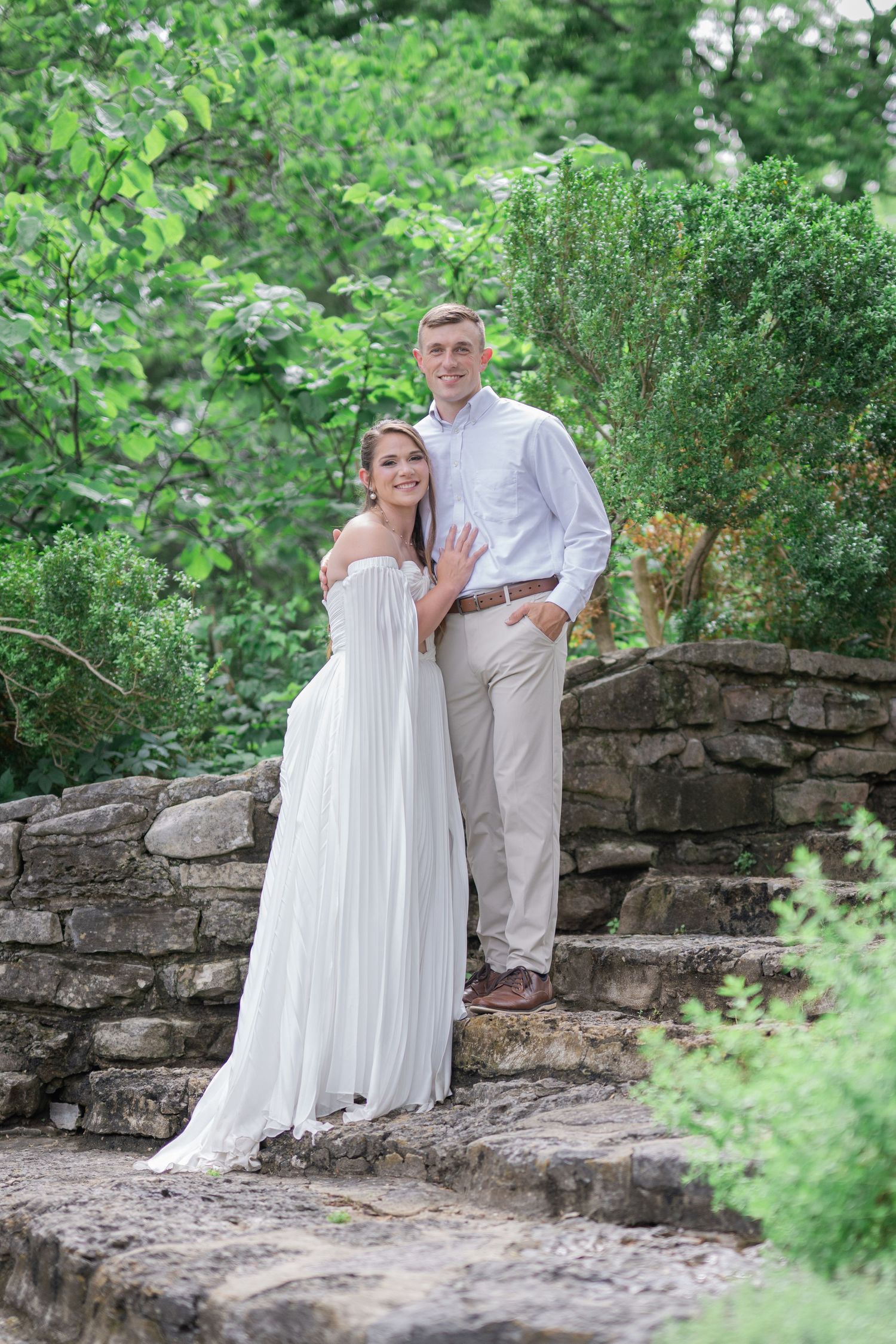 Couple in white dress and light blue shirt posing on stone steps surrounded by lush green trees.