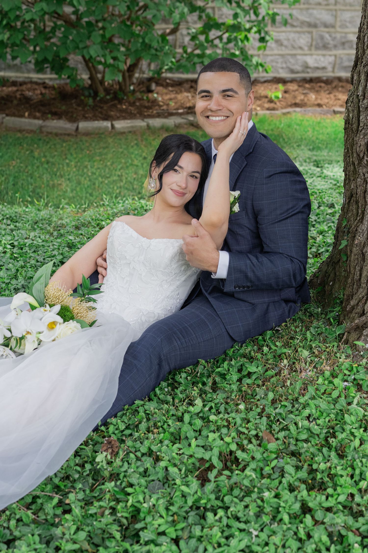 A smiling bride and groom sit together under a tree on green ground cover, the bride holding a white bouquet in a lace gown.