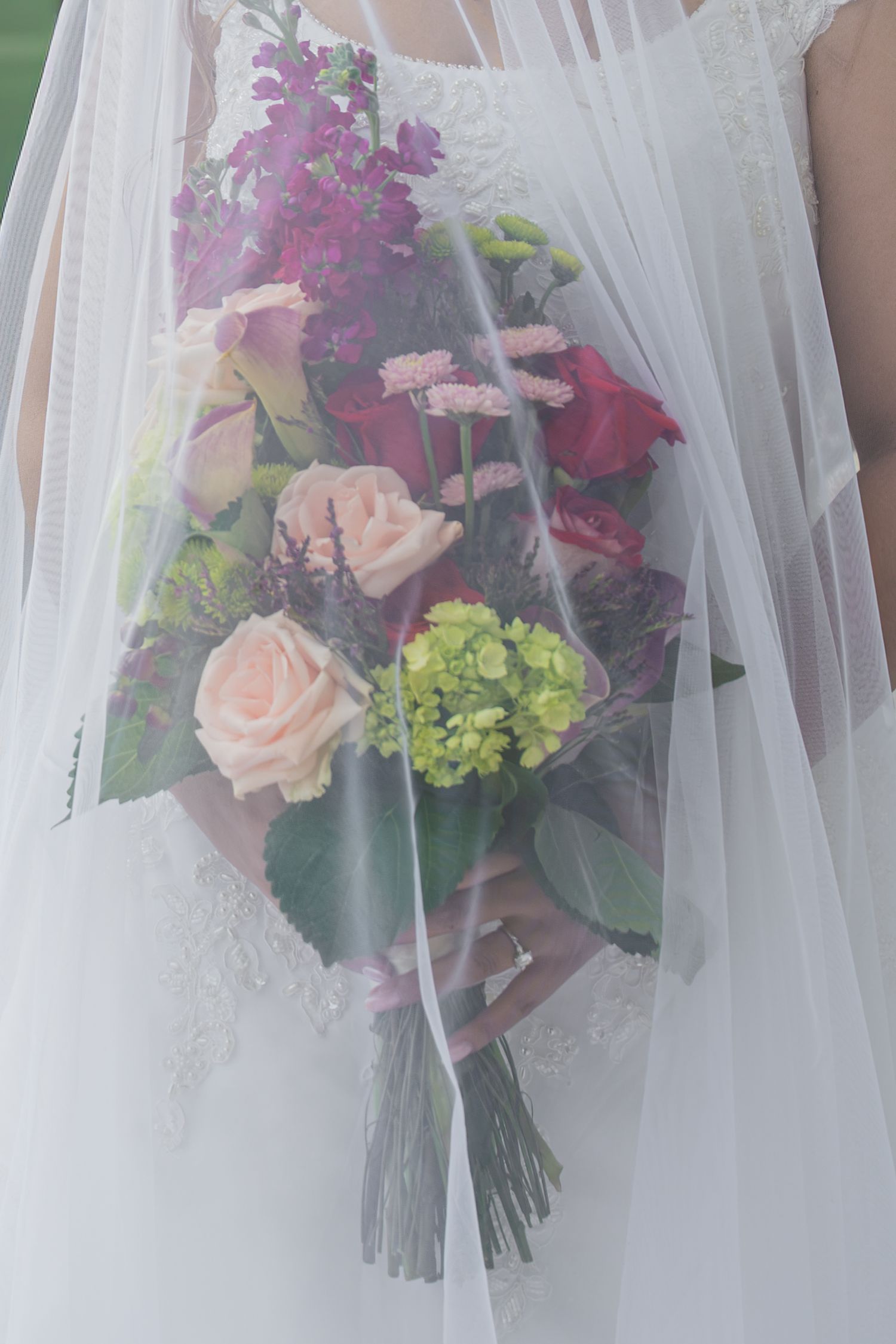 Bride holding a colorful wedding bouquet with pink roses, green hydrangeas, and purple flowers, draped with a white veil.