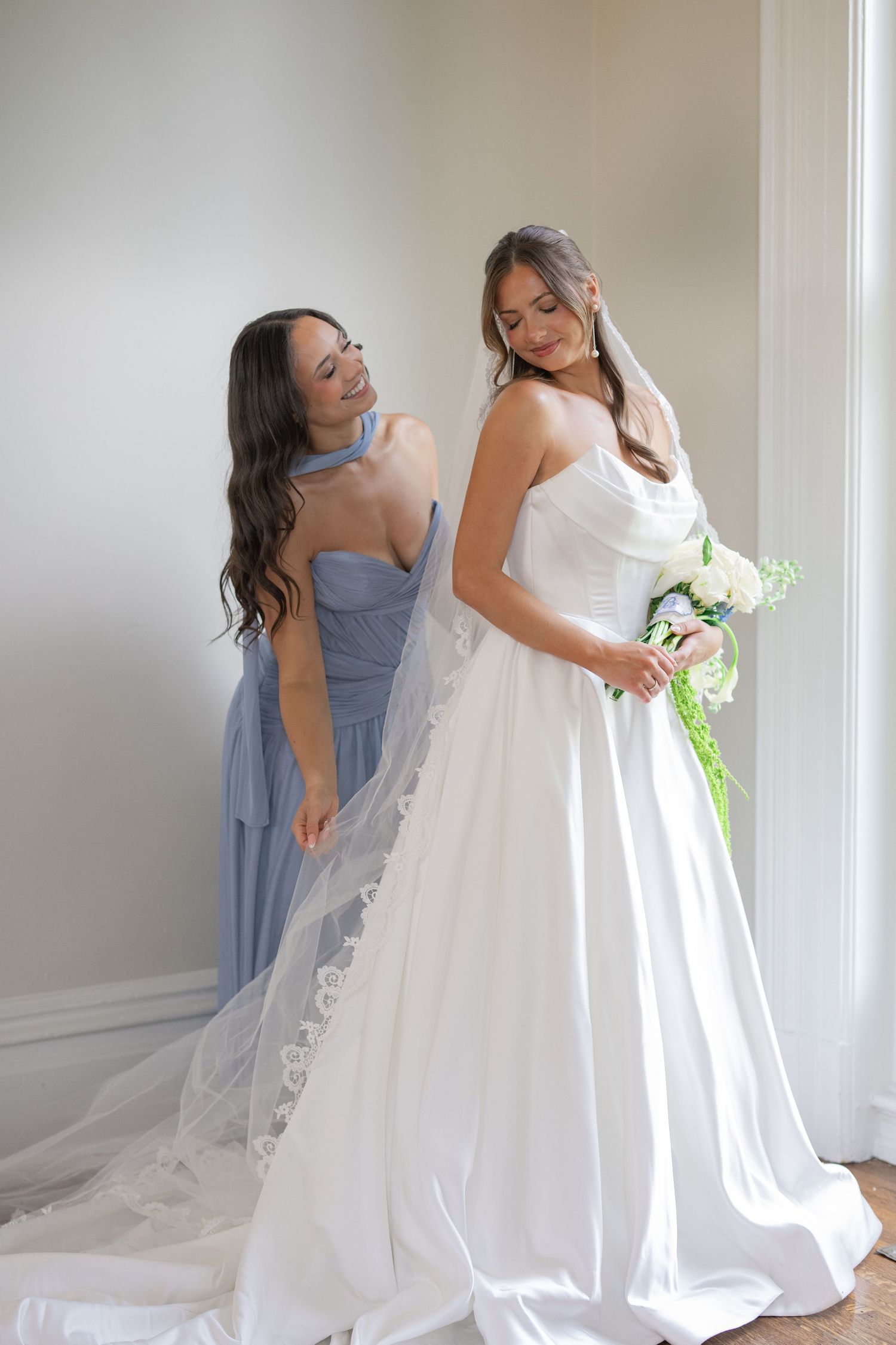 Bridesmaid in blue dress helping bride in white gown with veil while holding bouquet.