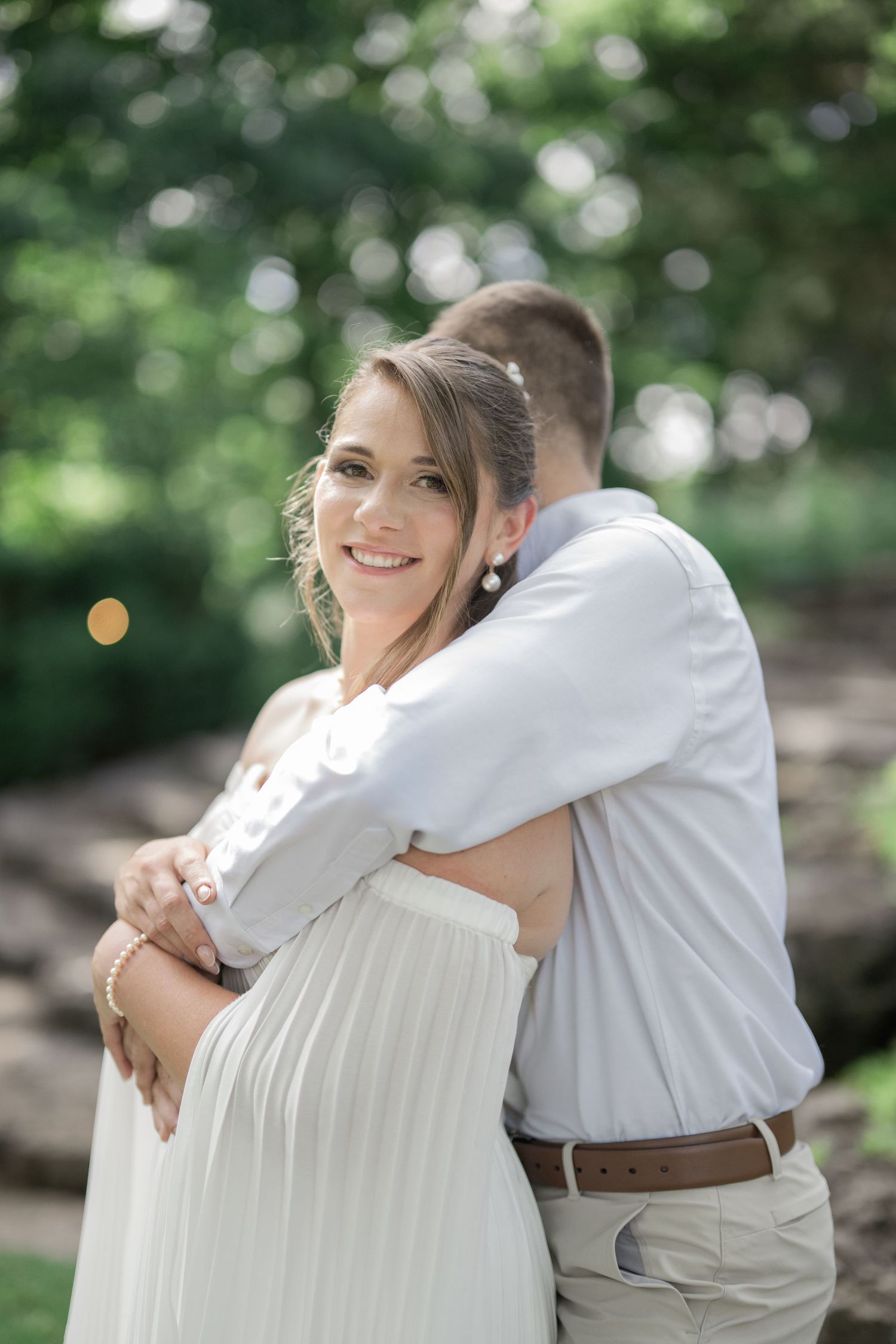 Pregnant woman in white dress smiles while partner embraces her from behind in a lush green outdoor setting.