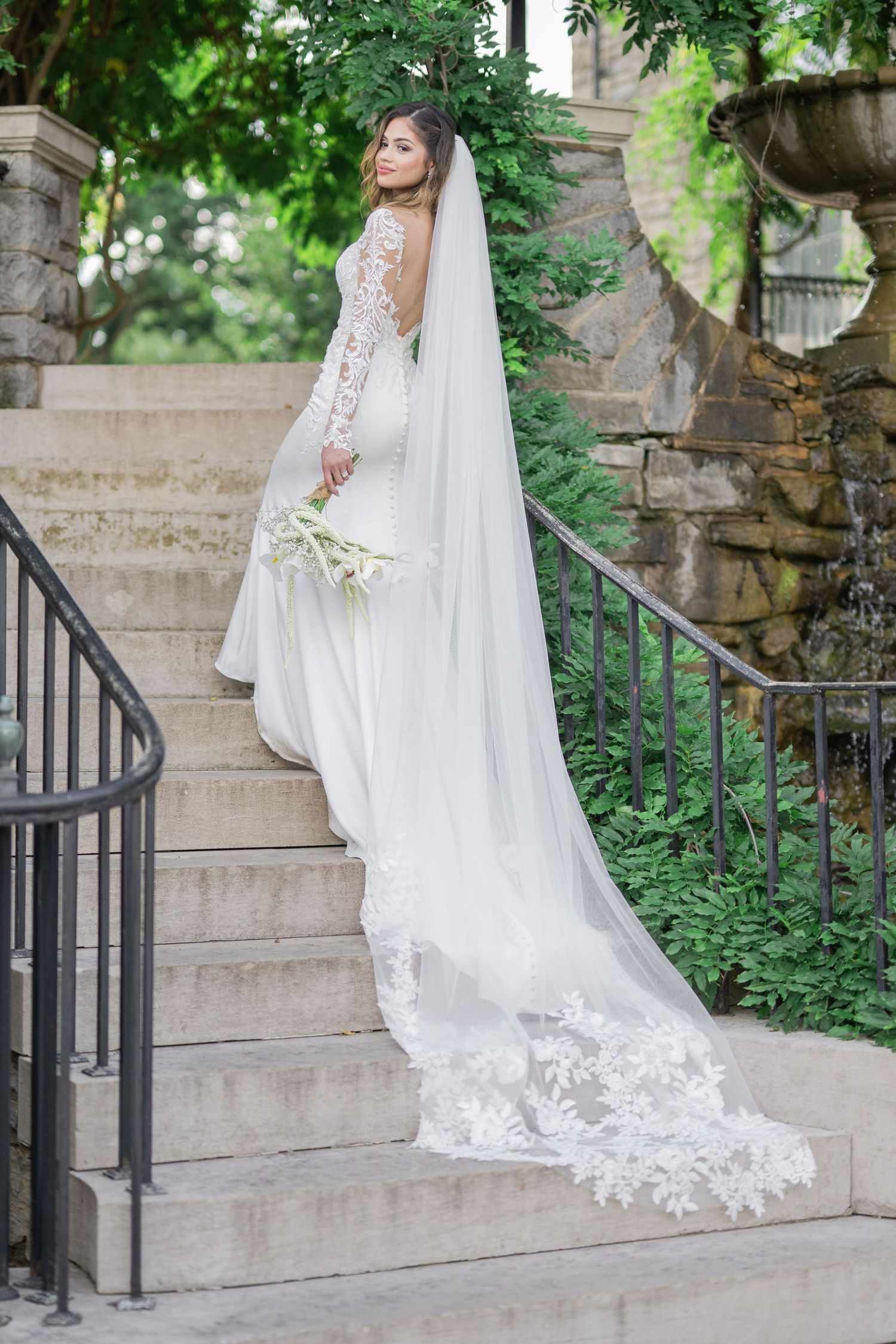 Bride in lace long-sleeve wedding gown with cathedral veil descending stone steps surrounded by lush greenery.