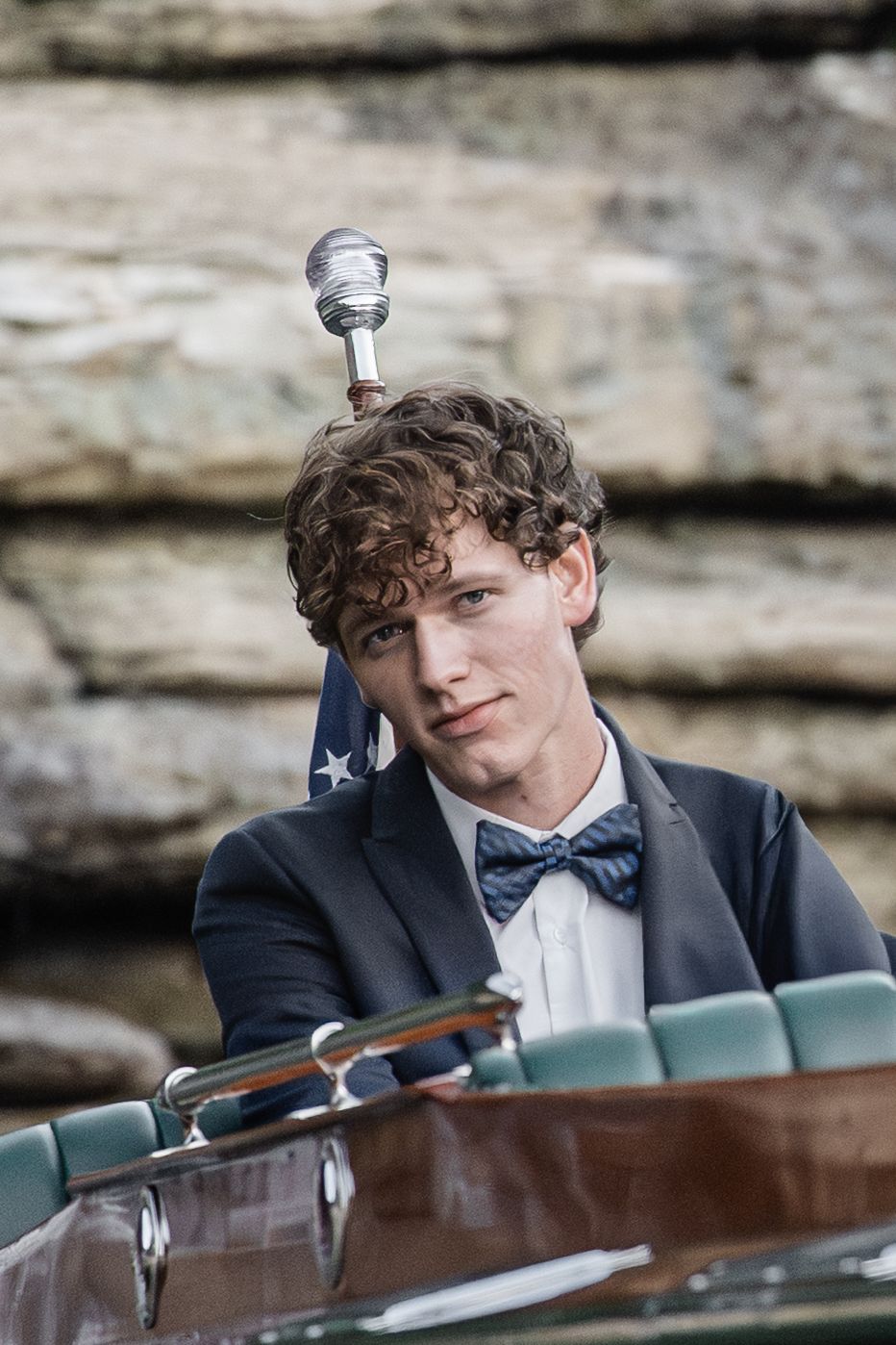 Young man in suit and bow tie sits at a piano podium with microphone, performing outdoors near a stone wall.
