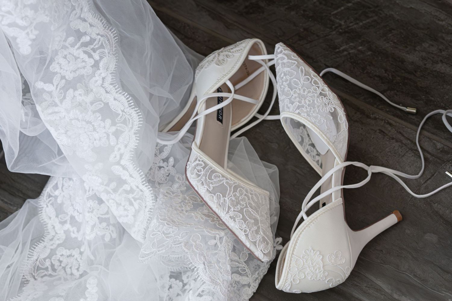 Elegant white lace bridal heels with strappy ankle ties displayed beside a flowing wedding veil on dark wood.