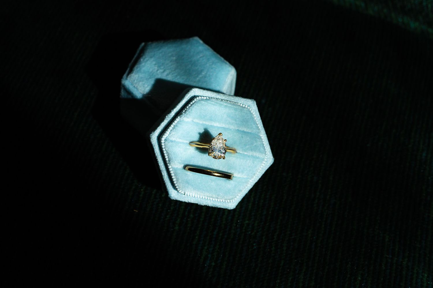 Gold diamond engagement ring displayed in a light blue hexagonal velvet ring box on dark fabric.