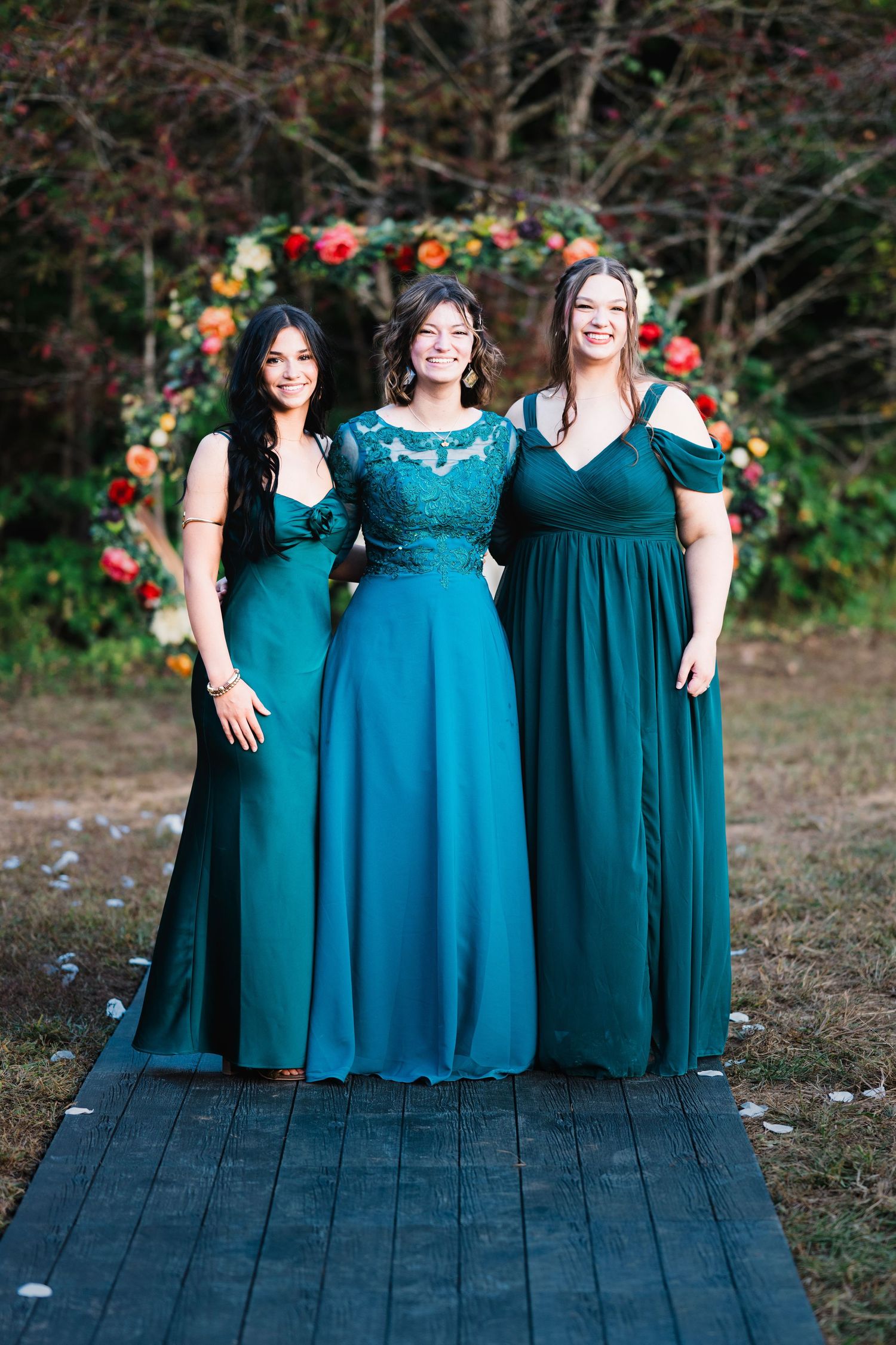 Three bridesmaids in teal green gowns smiling before colorful floral arch at outdoor wedding.