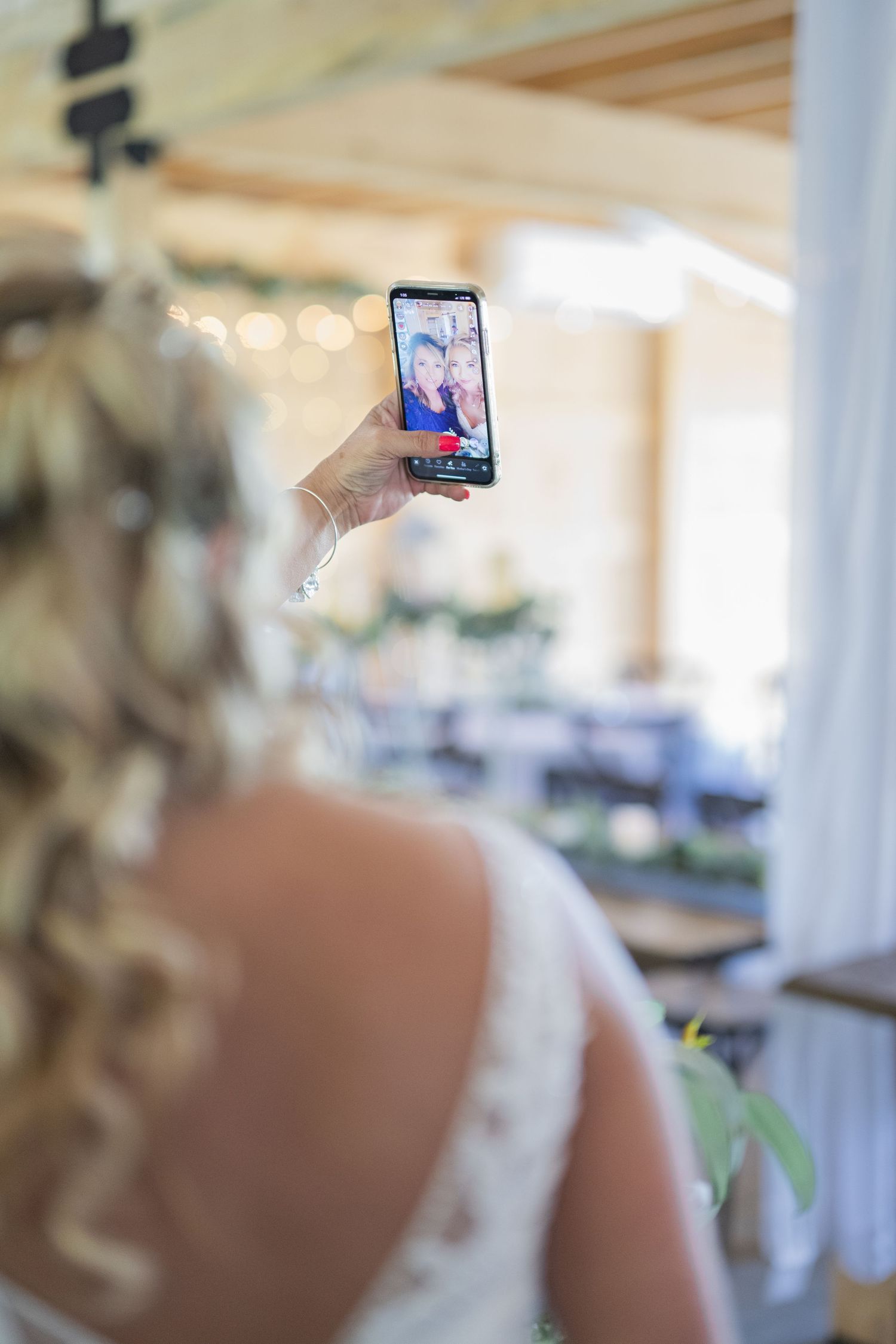Bride in lace dress holds phone showing FaceTime call with two smiling women at elegant wedding venue.