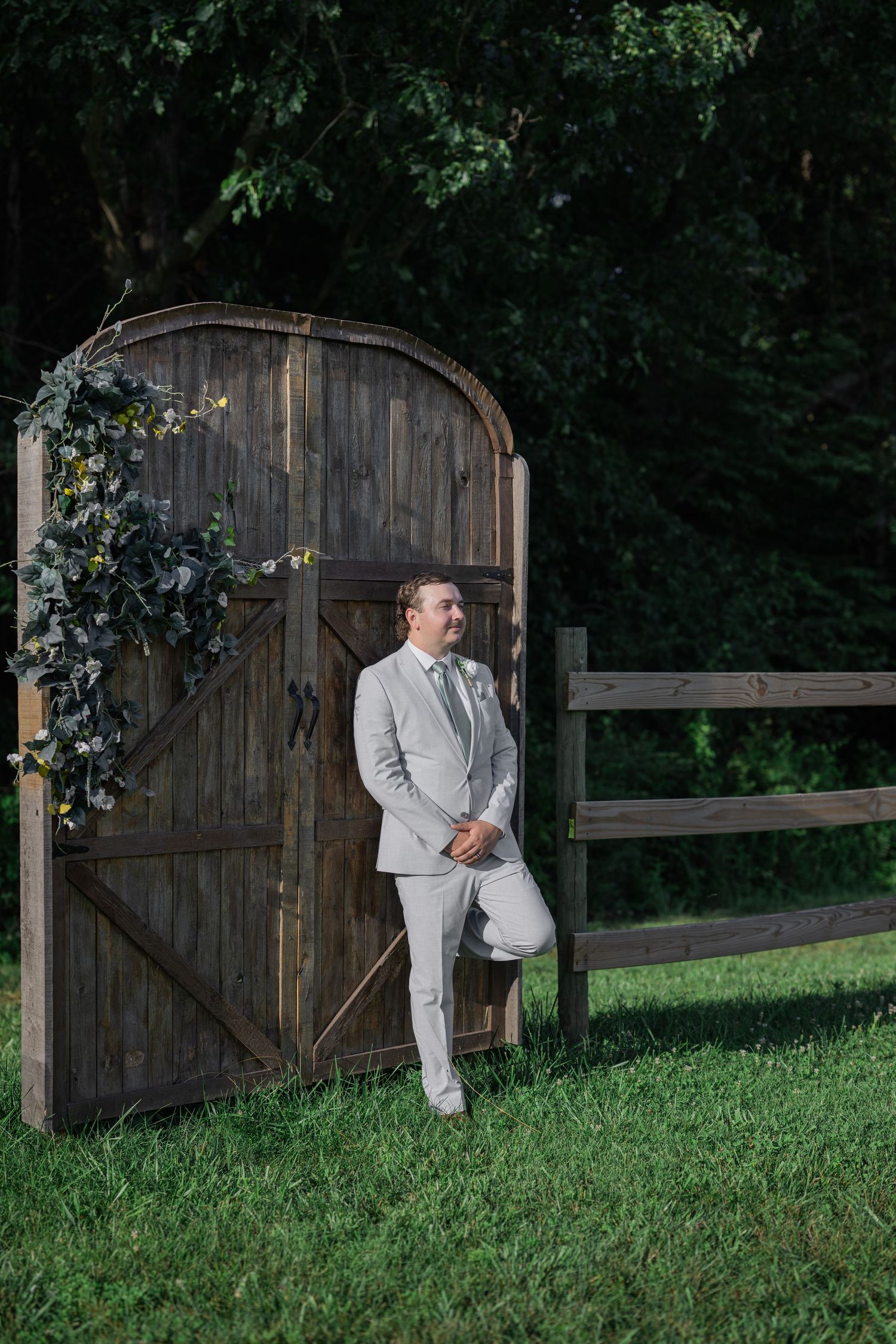 Groom in light gray suit leaning against rustic wooden gate with floral wreath at outdoor wedding venue.