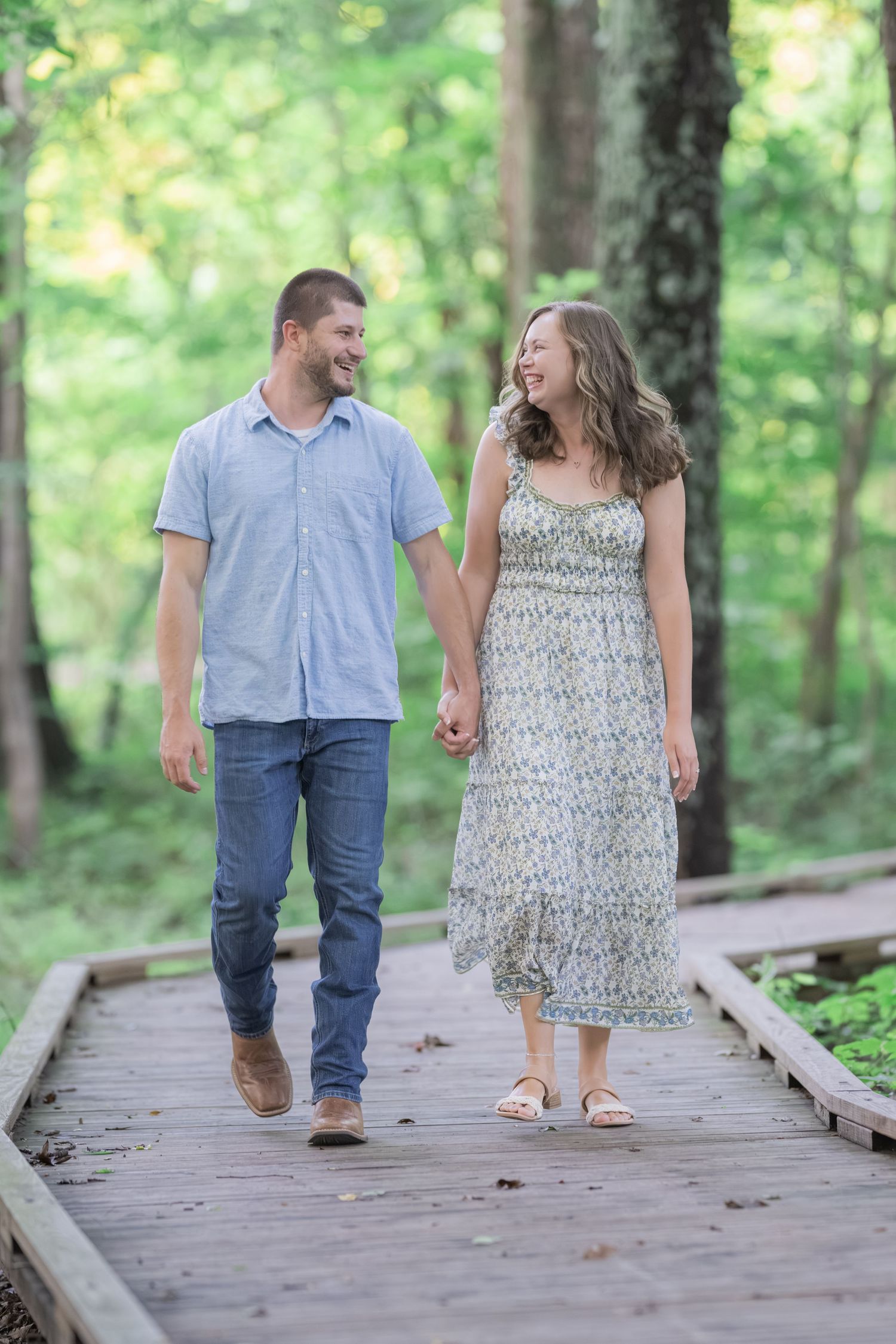 A smiling couple holds hands while walking on a wooden boardwalk path surrounded by lush green trees.