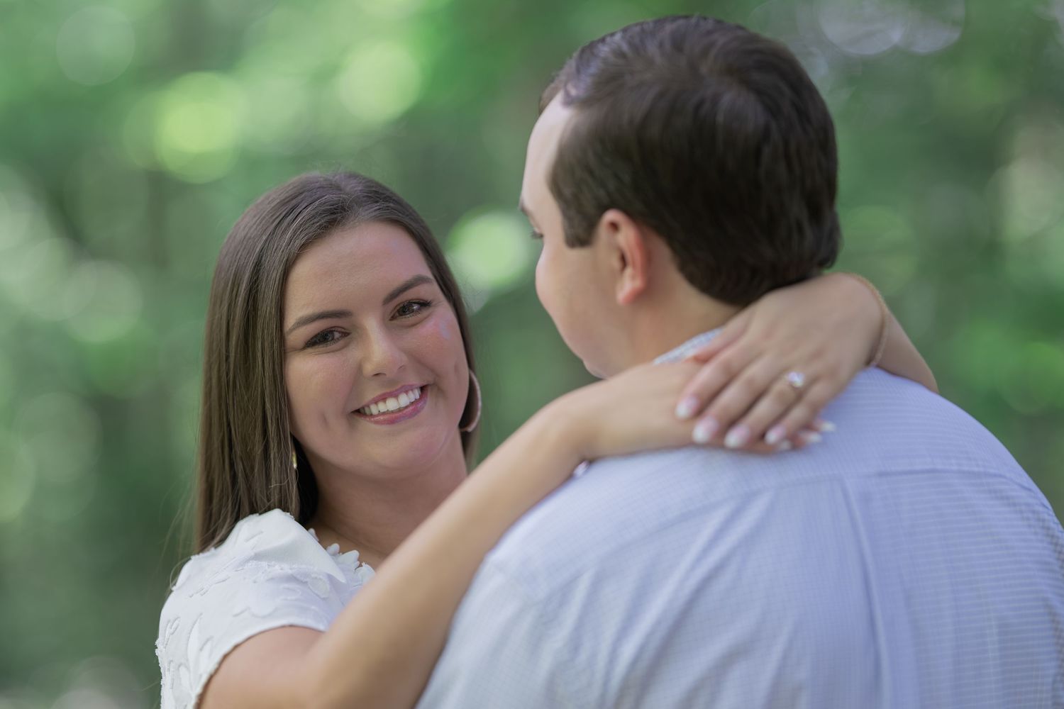 A smiling woman in a white dress embraces a man outdoors, with lush green trees in the background.