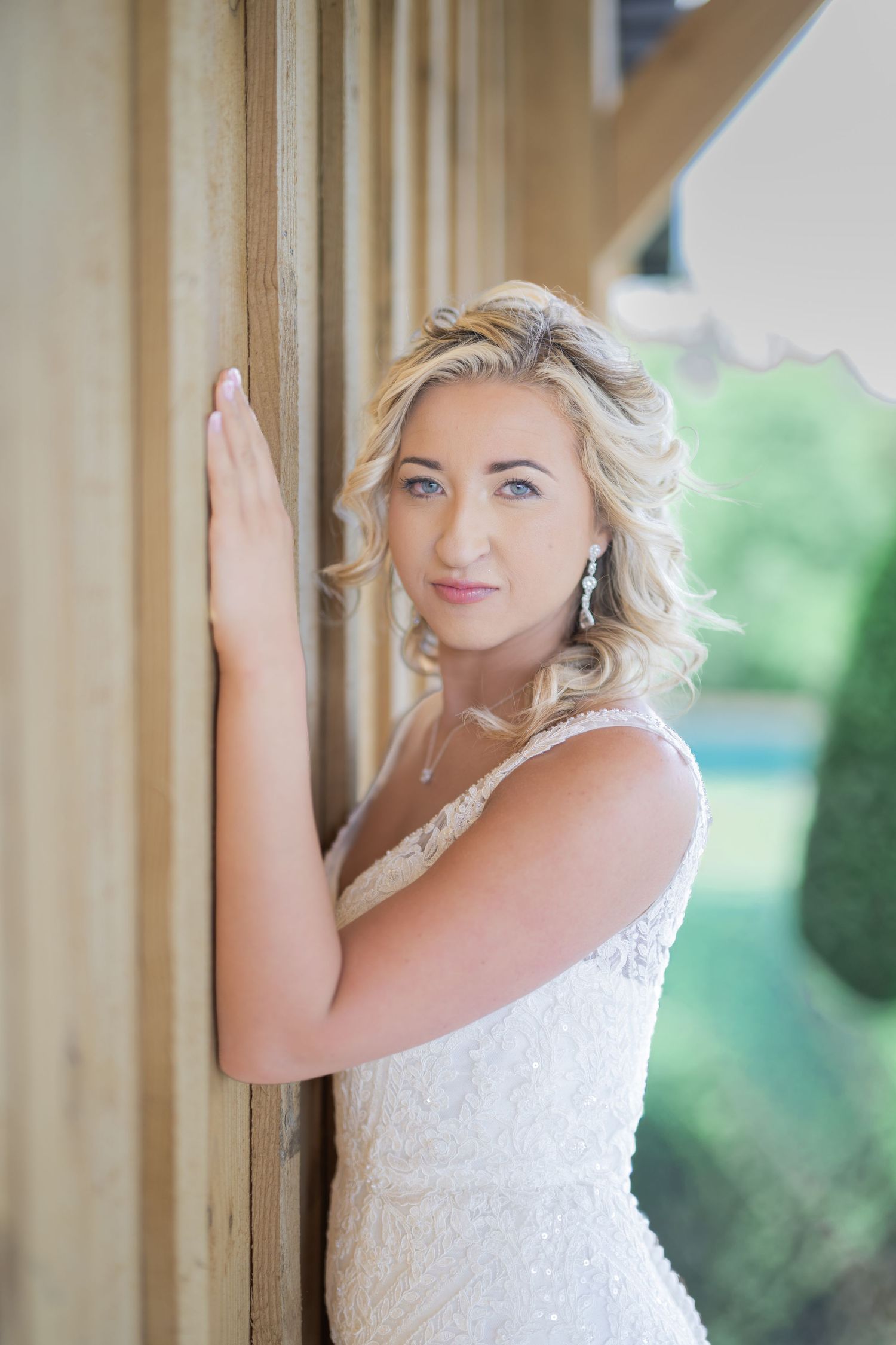 Blonde bride in white lace gown leaning against wooden column, wearing drop earrings, with soft curled hair.