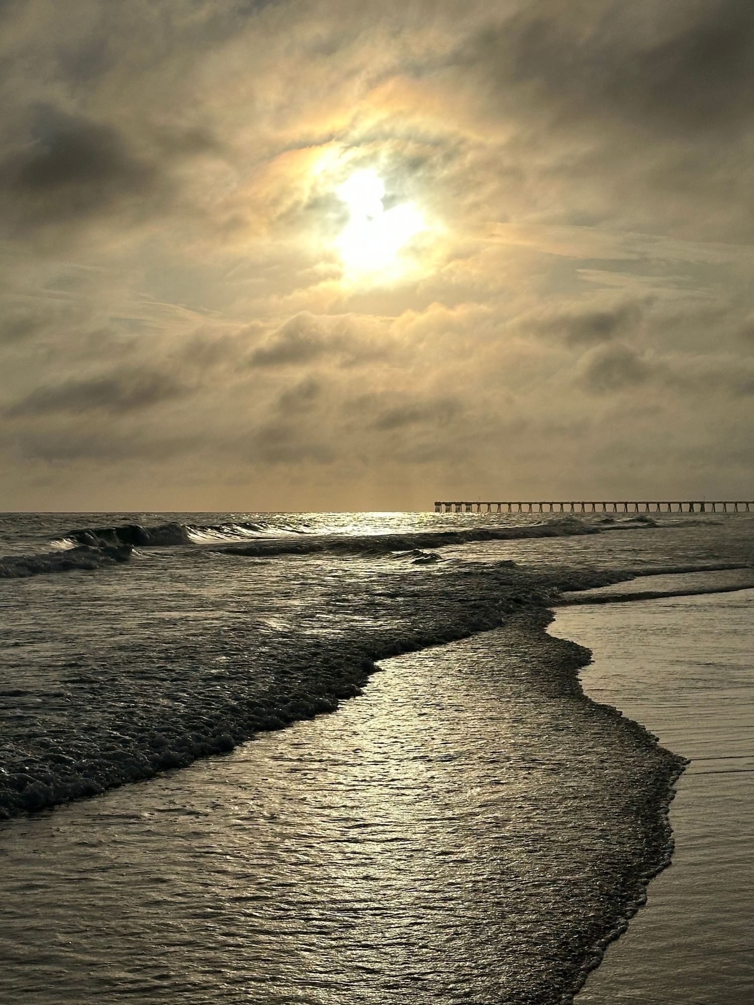 Dramatic beach sunset with golden sun breaking through cloudy sky, reflecting on ocean waves near a distant pier.