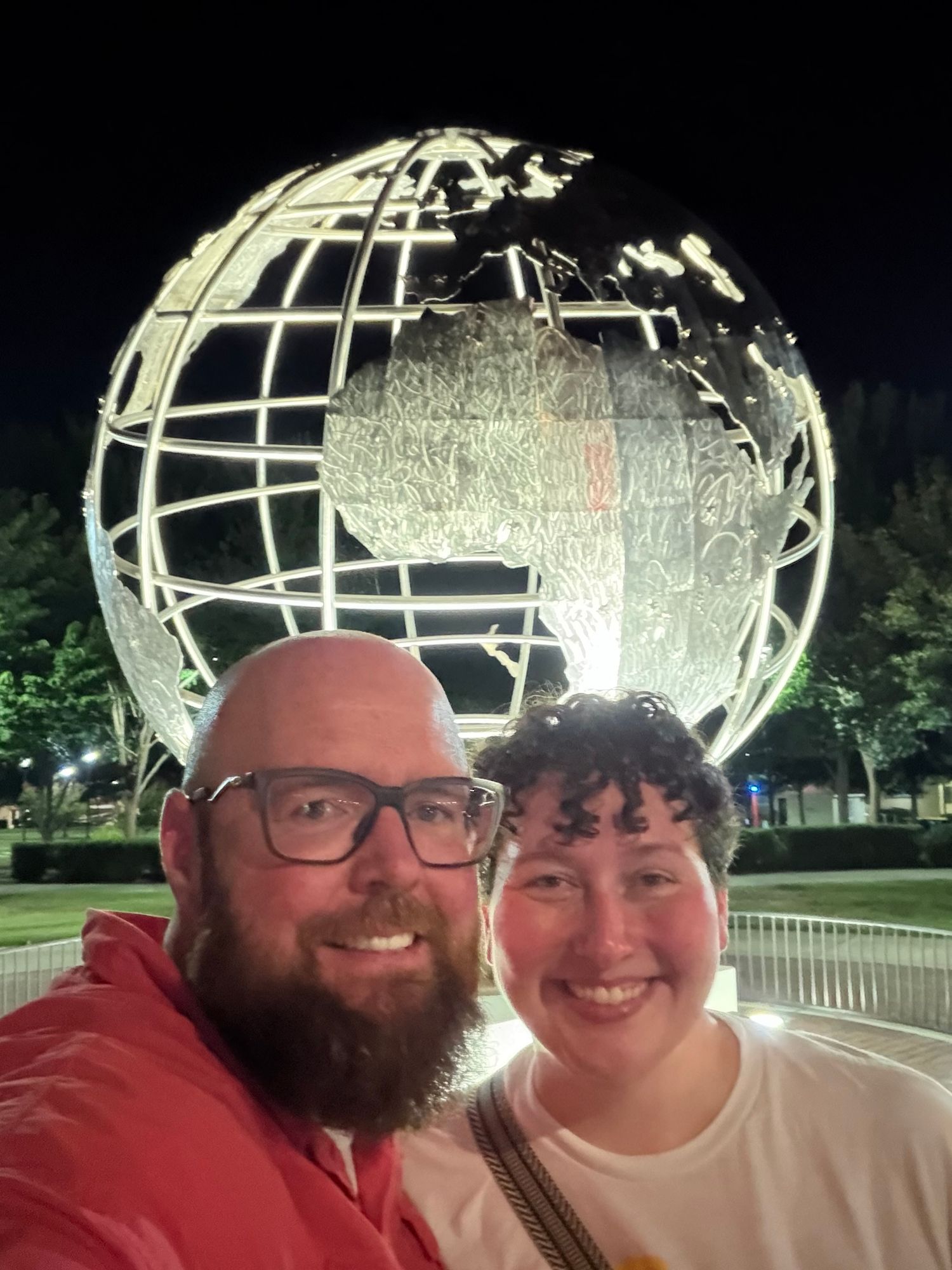 Couple smiling in selfie at night in front of illuminated Unisphere globe sculpture at Flushing Meadows Park, NYC.
