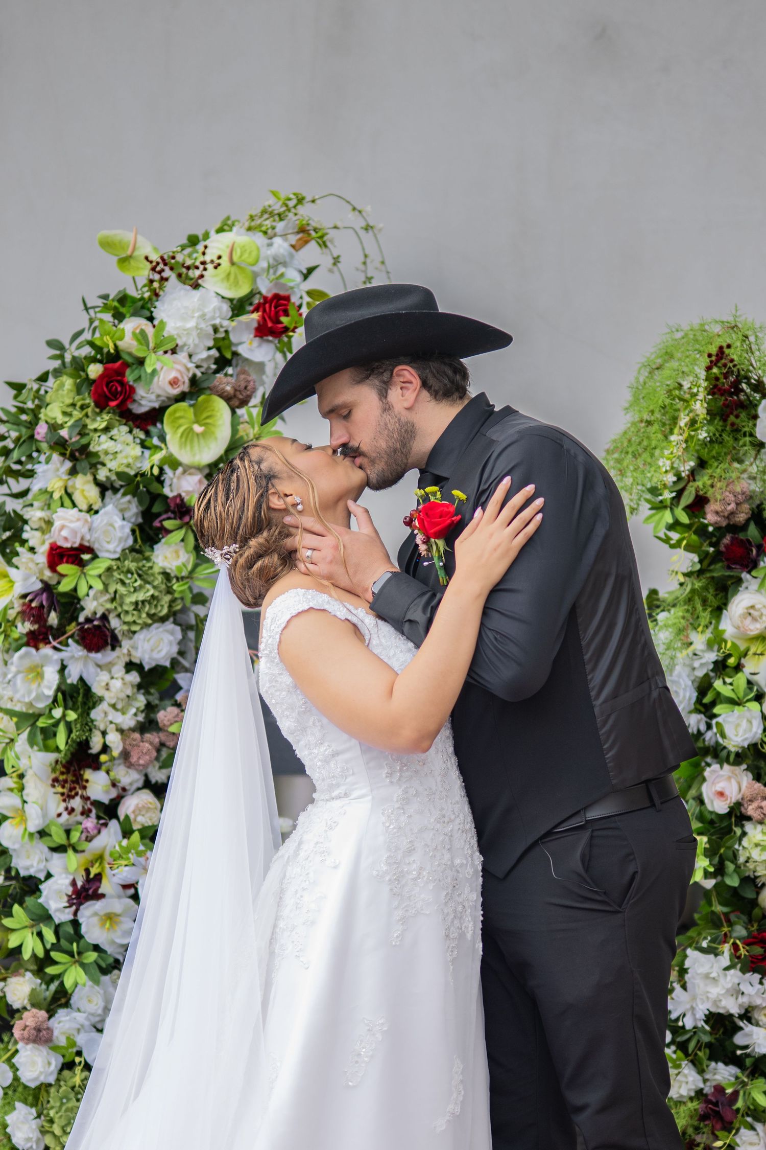 Bride and groom share first kiss at floral arch, groom wearing black cowboy hat and suit, bride in white lace gown.