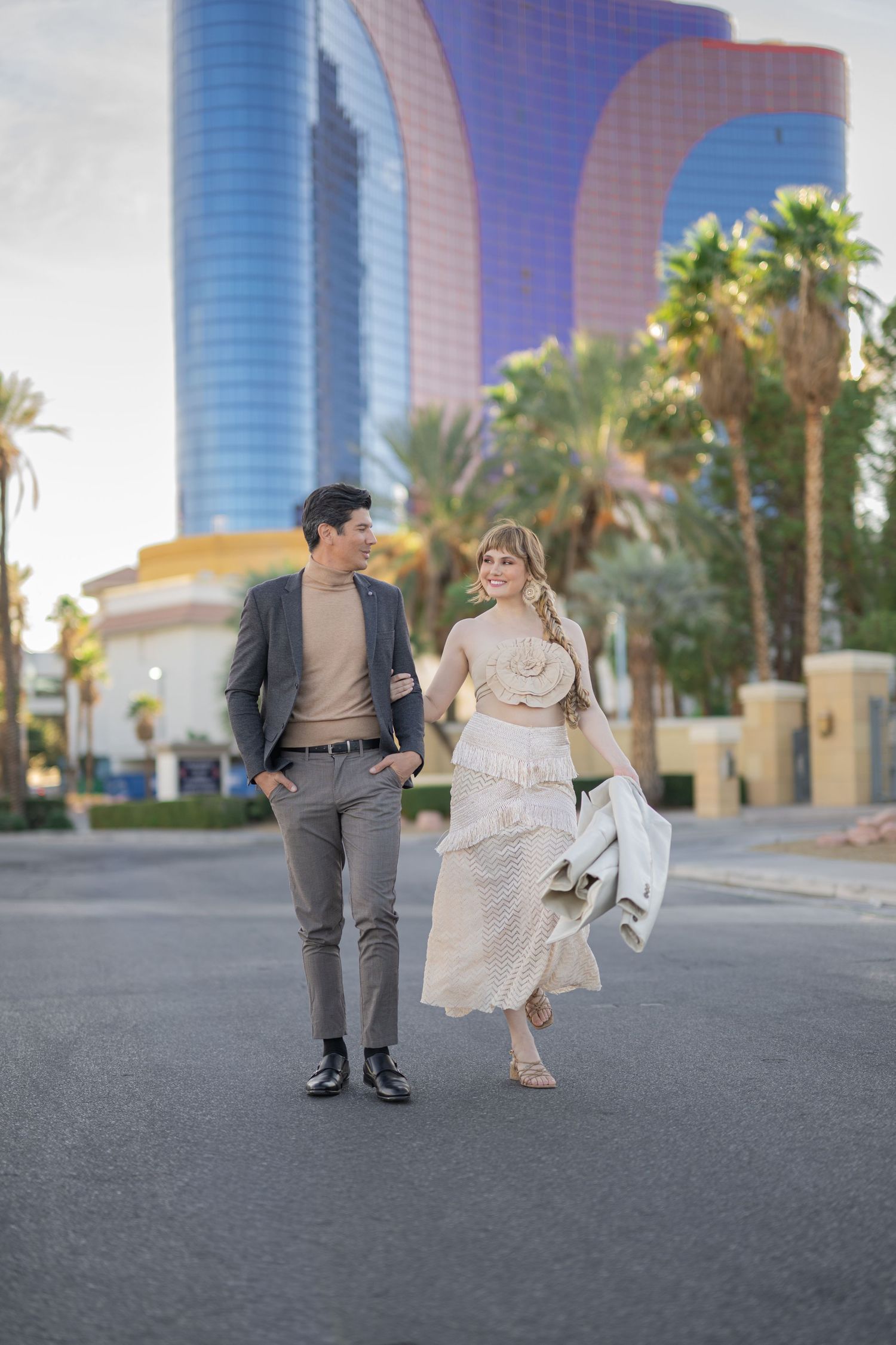 Elegant couple walking outdoors near modern glass tower and palm trees, woman in flowing dress, man in gray suit.