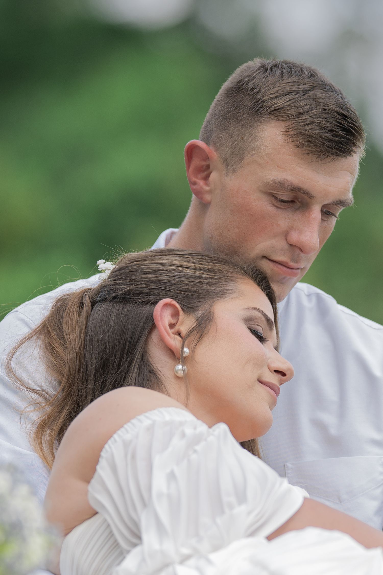 Romantic wedding couple in white attire embrace outdoors, bride wearing pearl earrings with groom tenderly looking down.
