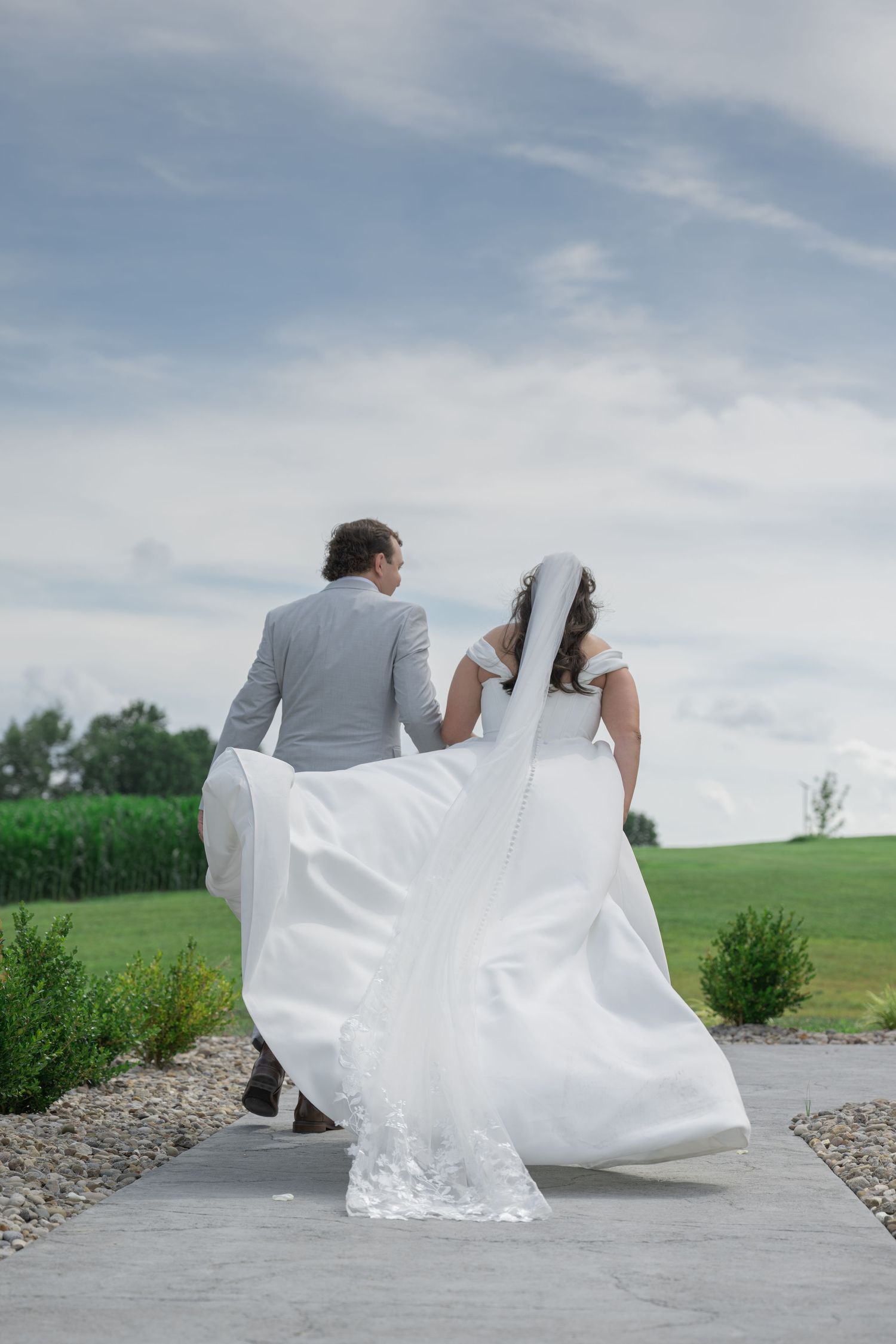 Bride and groom walking away on a path, white wedding dress flowing in the wind, green fields and cloudy sky.
