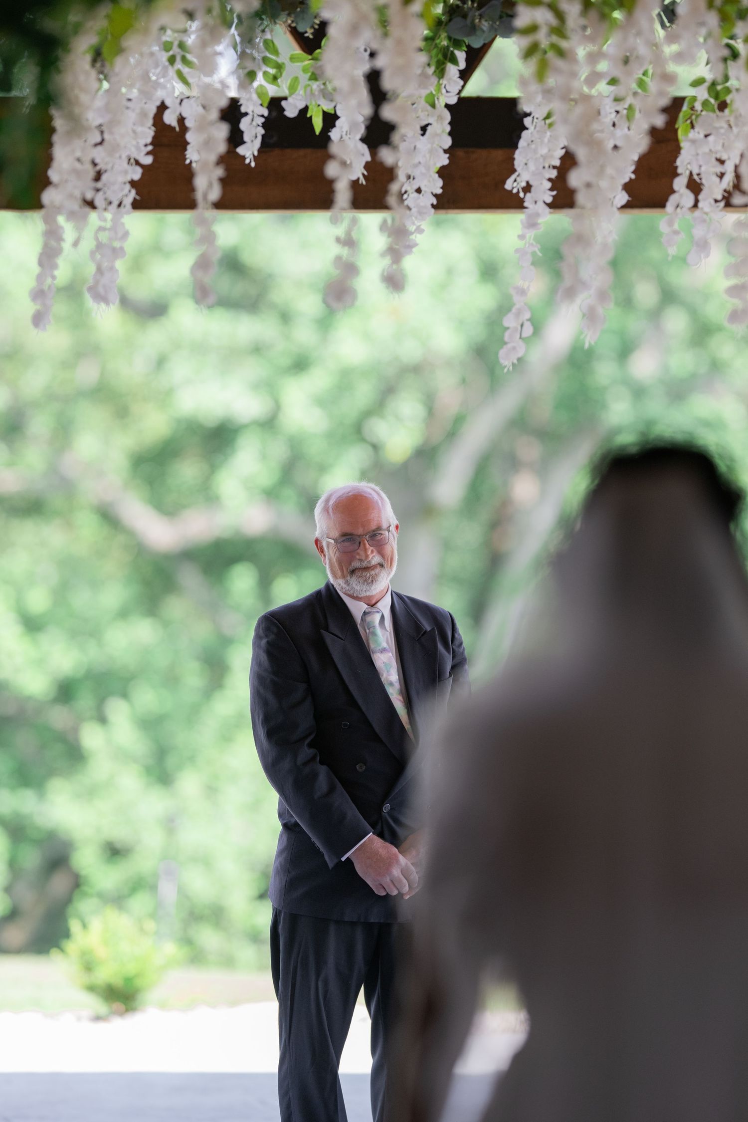 Elderly groom in black tuxedo awaits bride at outdoor wedding ceremony adorned with white wisteria flowers.