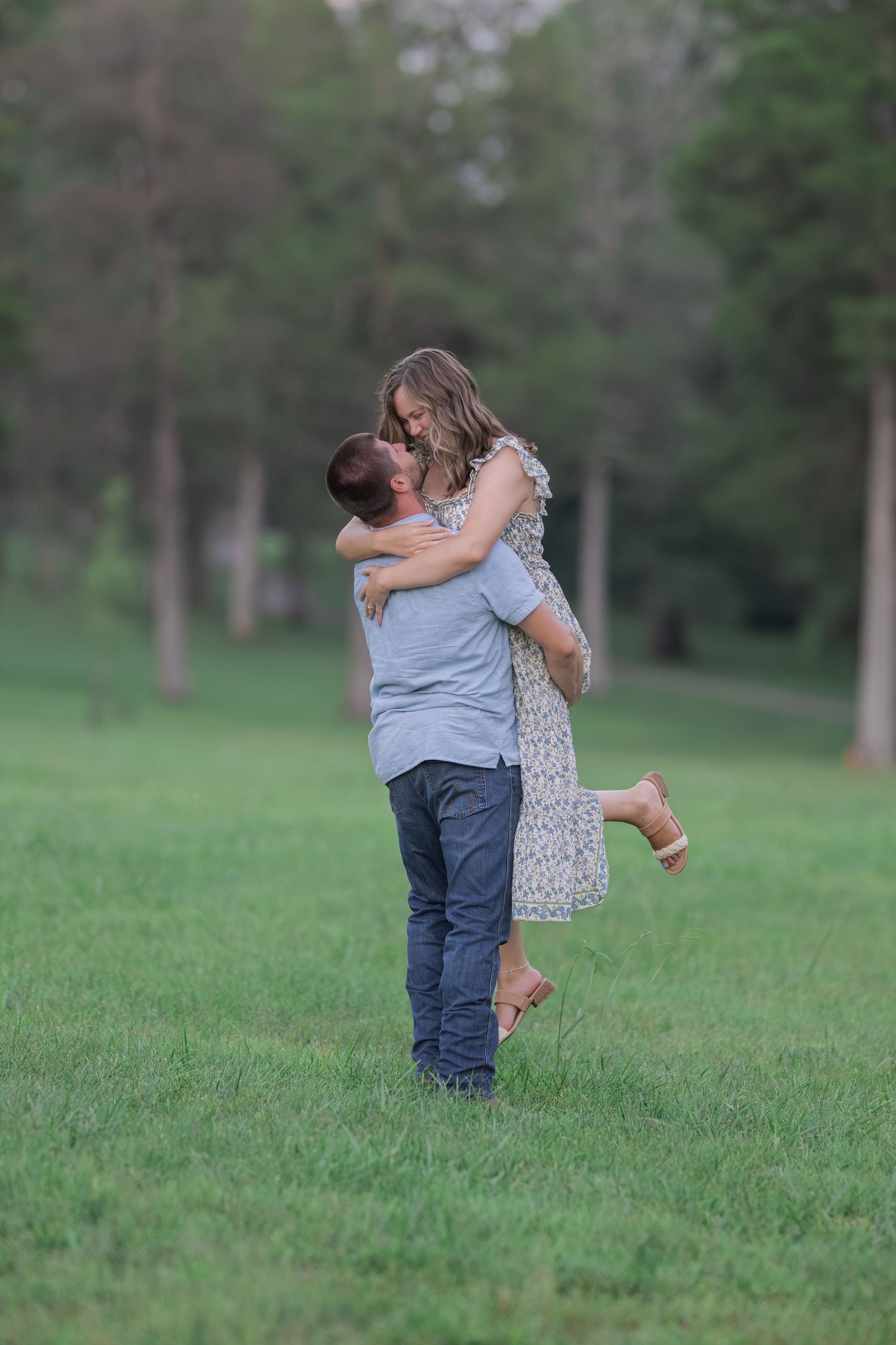 A romantic couple shares a kiss as the man lifts the woman in a green meadow surrounded by trees.
