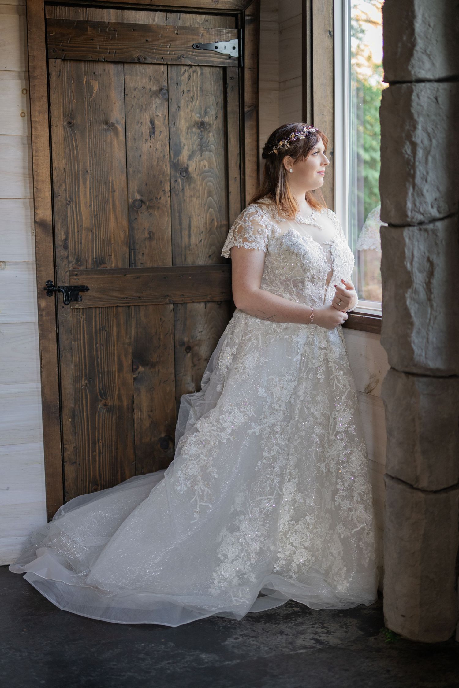 Bride in lace wedding gown with floral crown gazes out window beside rustic wooden barn door.