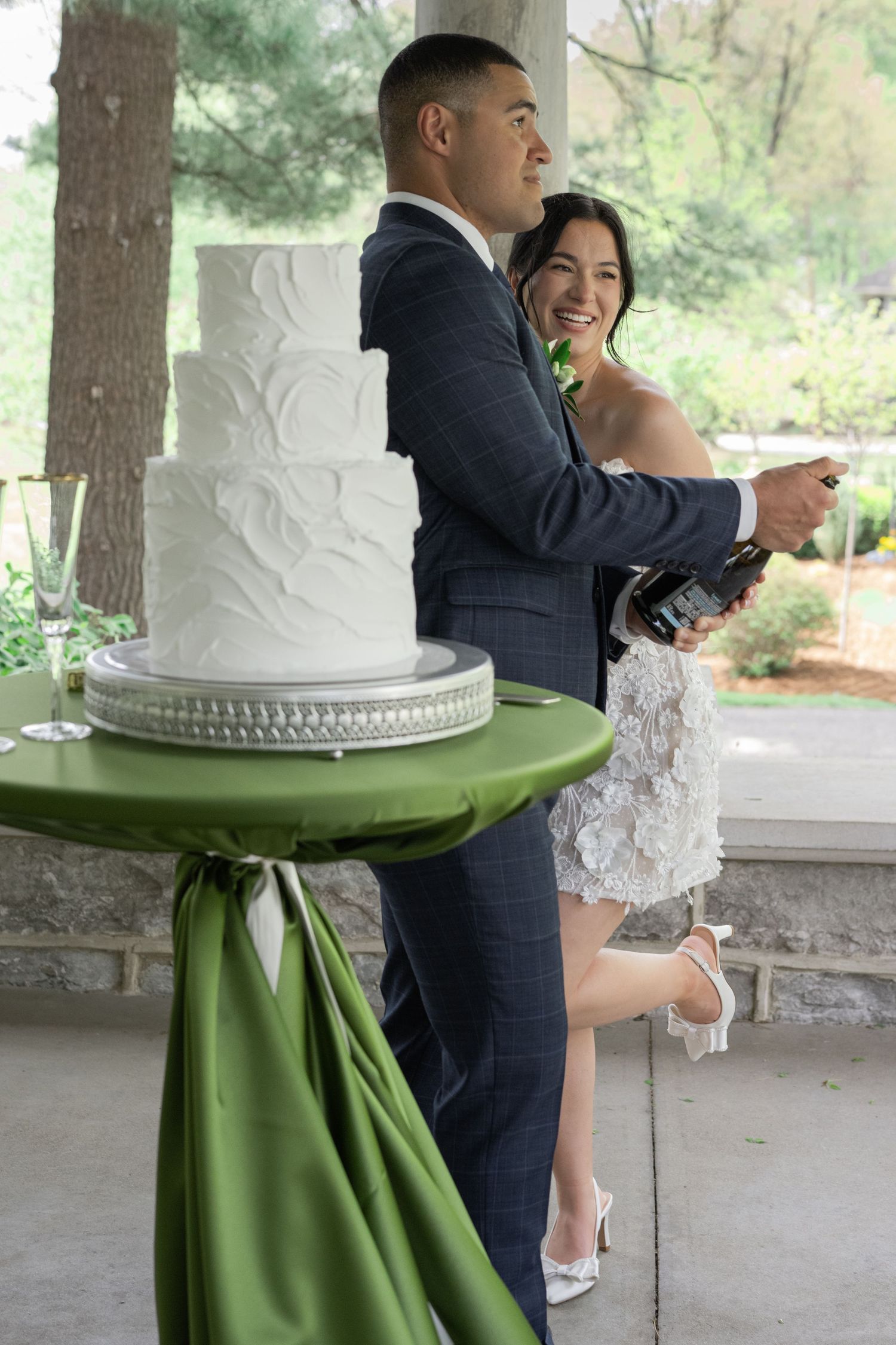 Bride and groom laugh while opening champagne beside a white tiered wedding cake on a green-draped table.