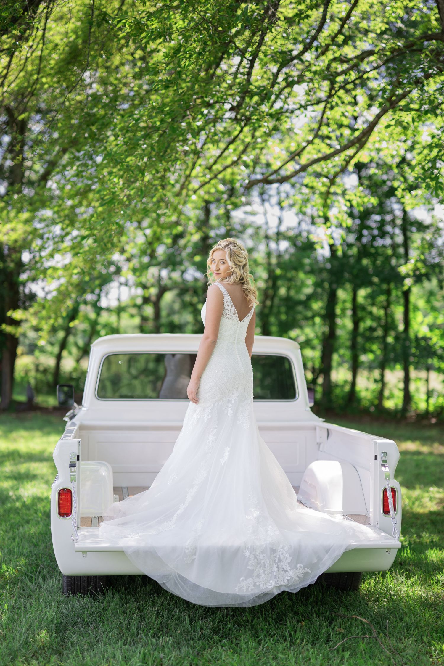 Bride in white lace gown stands in truck bed amid lush green trees, her elegant train draped over the tailgate.