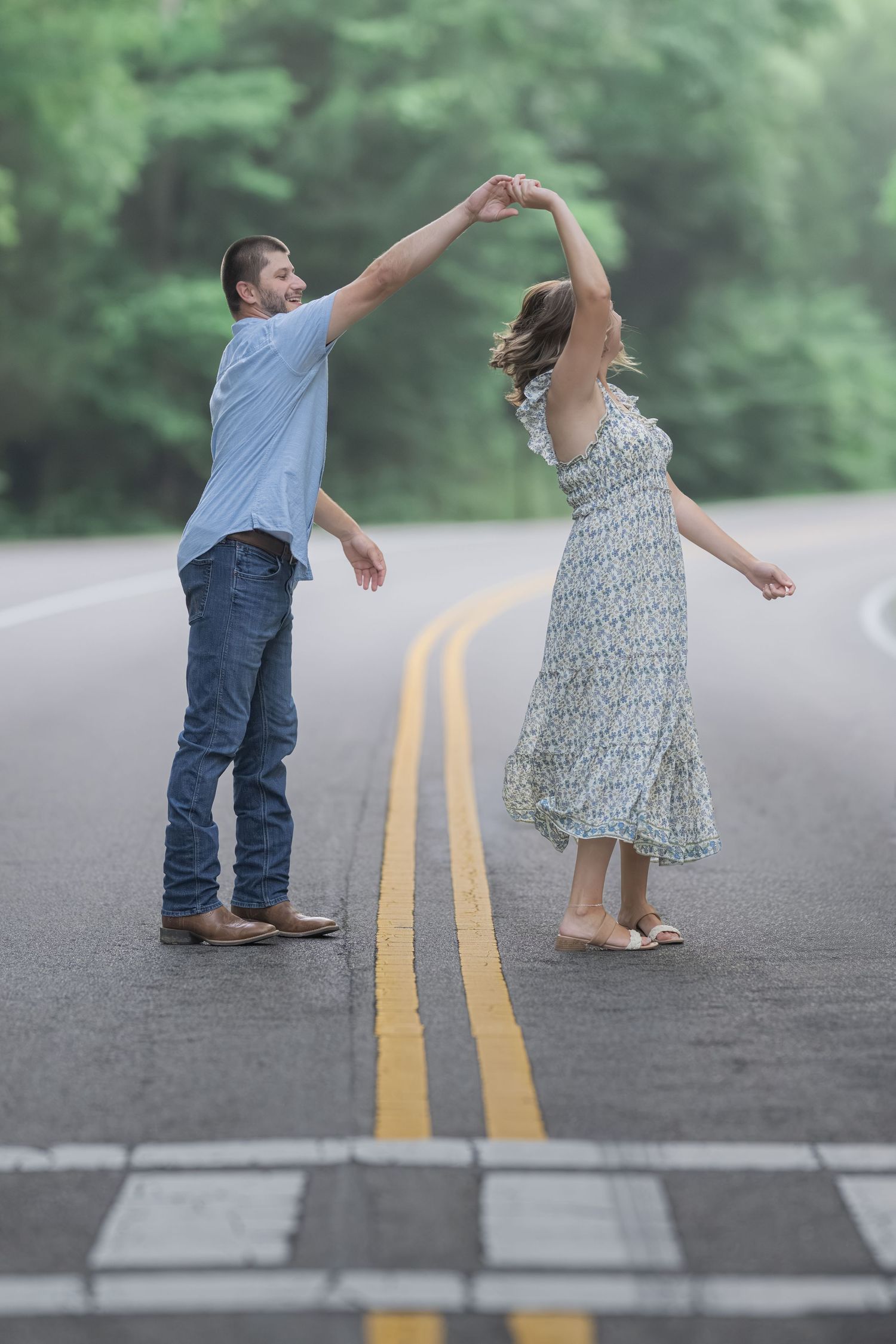 Couple dancing together on a road surrounded by lush green trees, man spinning woman in a floral dress.