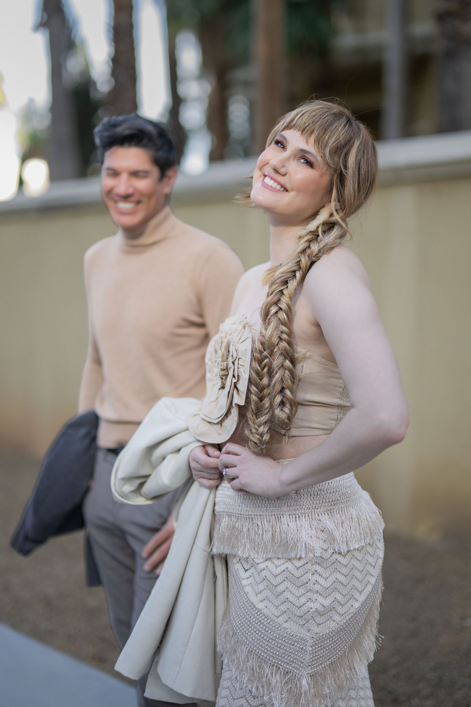 A smiling woman with a long braid in a cream dress holds a jacket, with a man in a beige turtleneck behind her.
