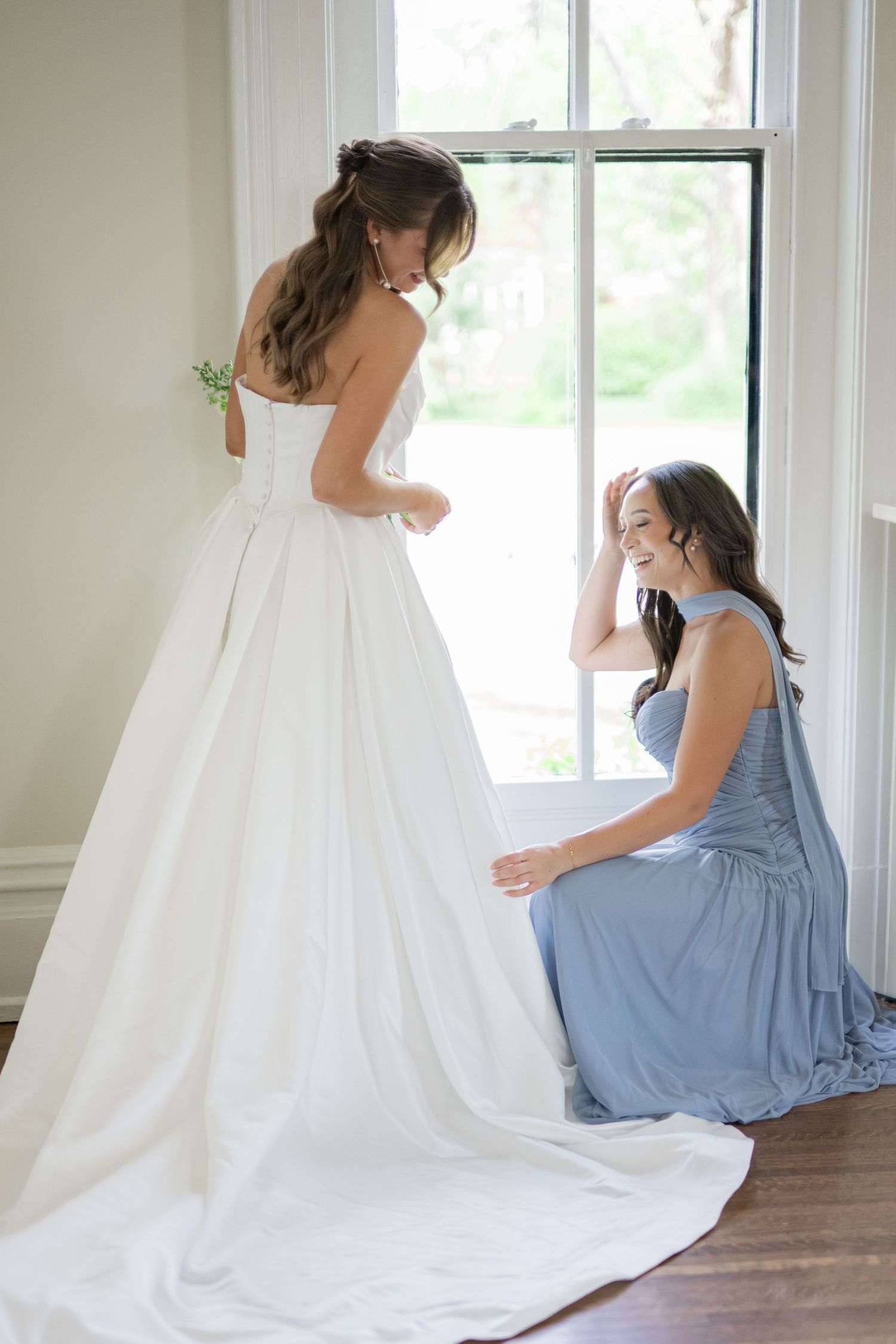 Bride in white gown stands by window as smiling bridesmaid in blue dress adjusts her train.