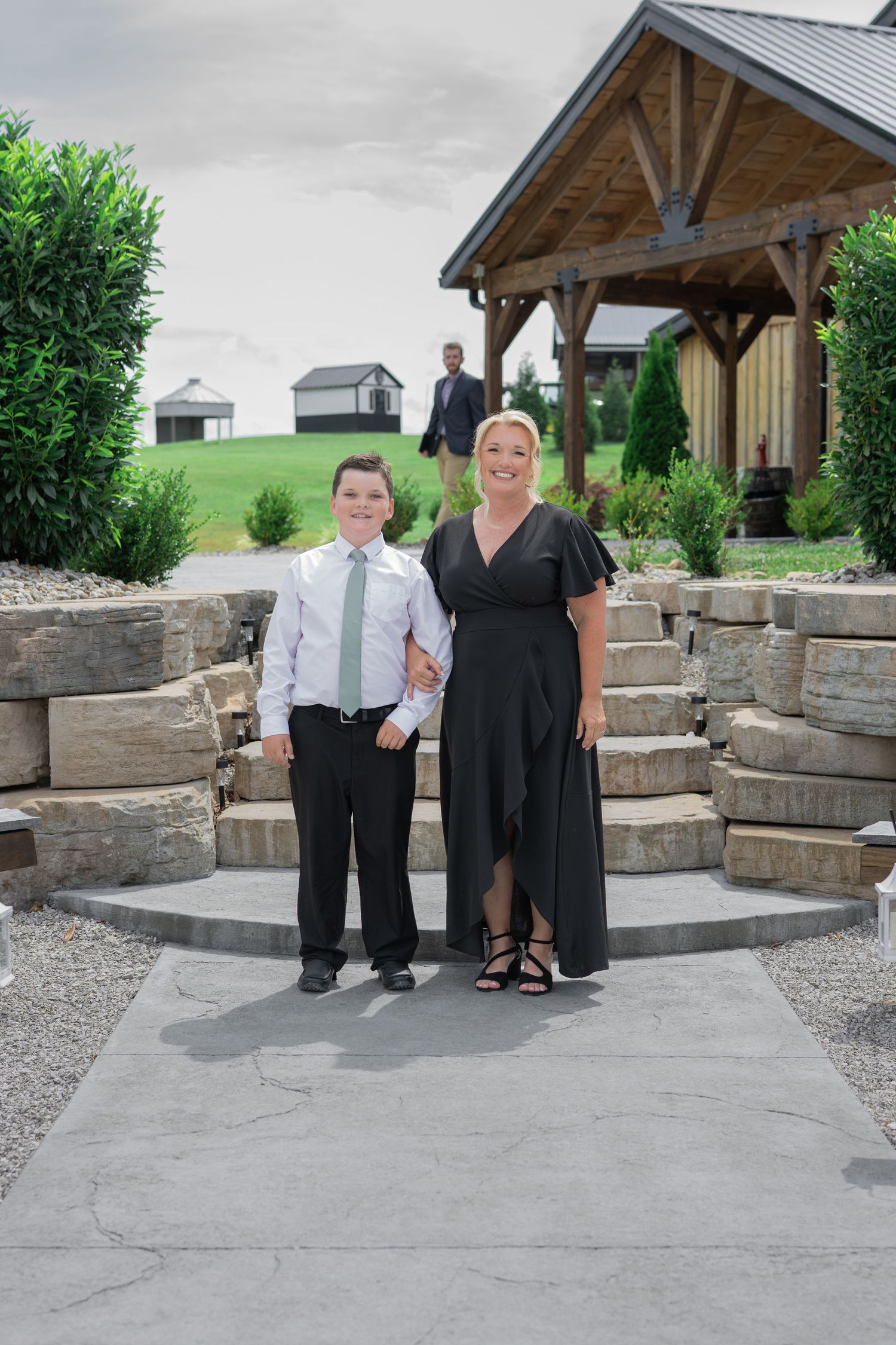 A woman in a black dress and a boy in a suit pose together at an outdoor wedding venue with stone steps and a wooden pavilion.