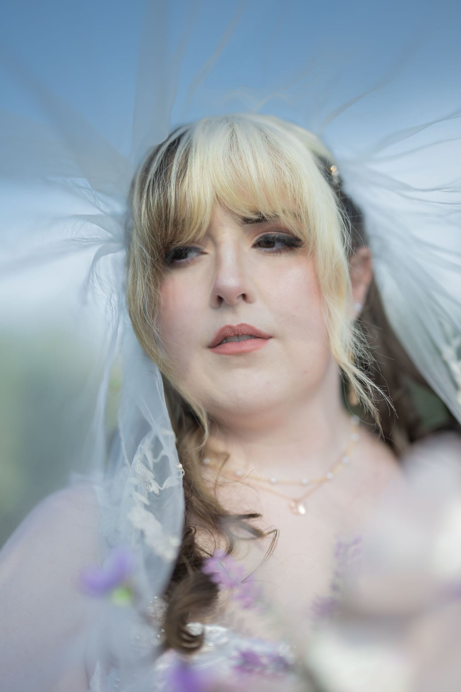 Bride in white veil and blonde hair with braids poses outdoors holding flowers.