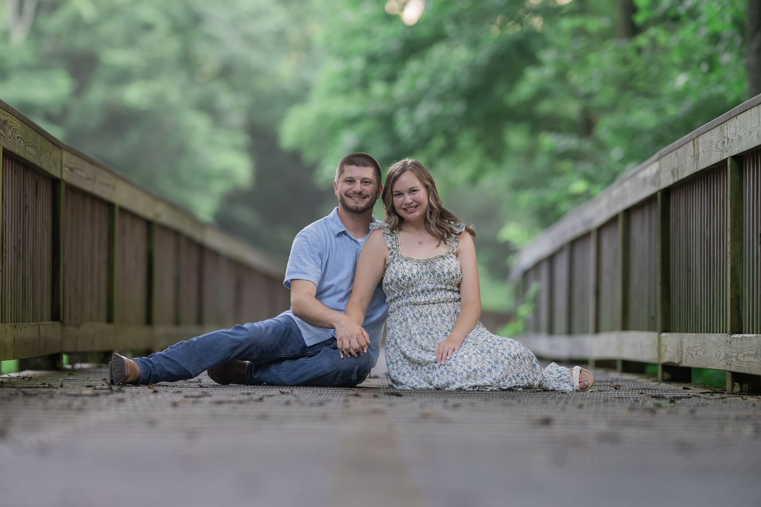 Couple sitting together on a wooden bridge surrounded by lush green trees for an outdoor engagement photo.