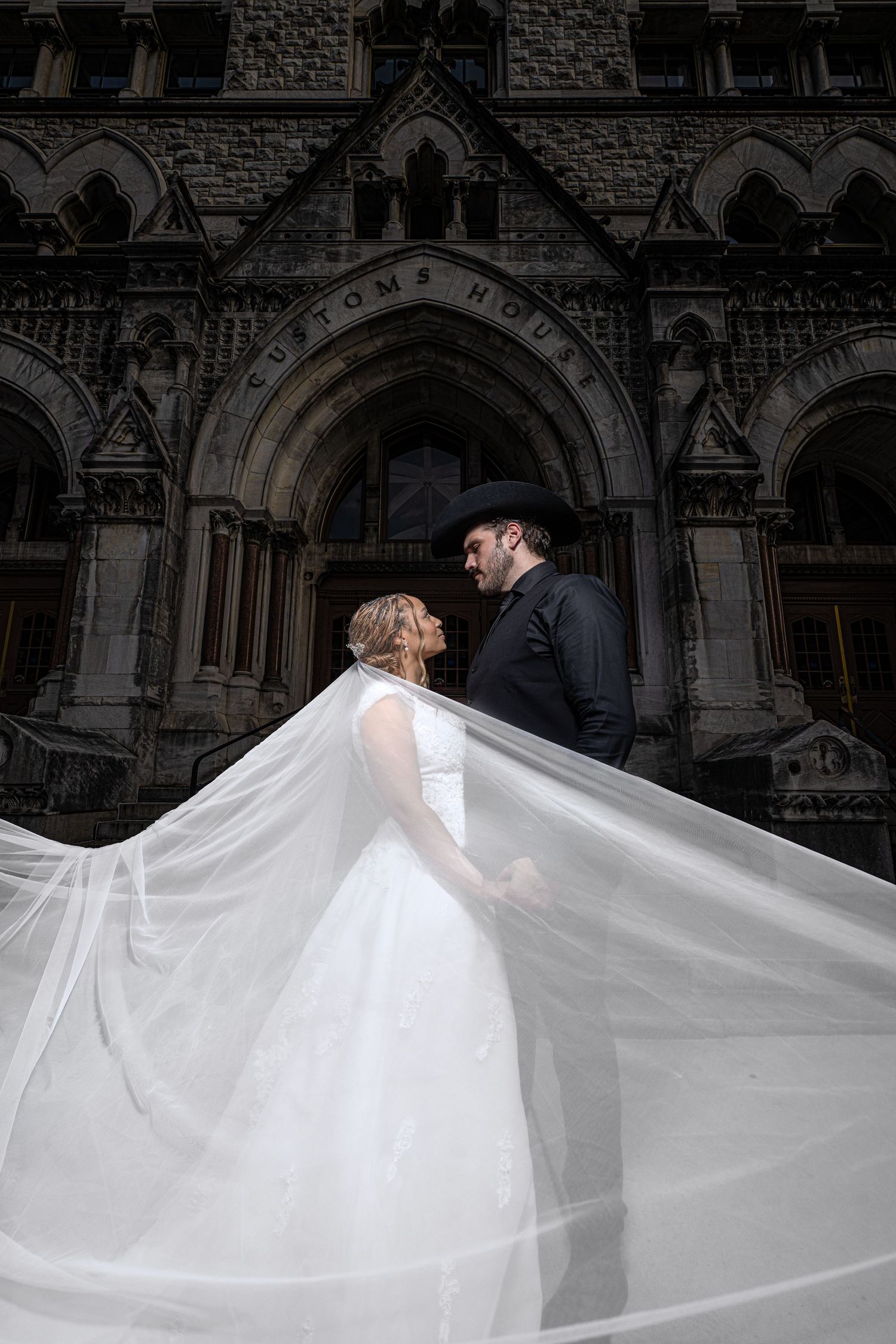 Bride and groom embrace before historic Customs House, her veil flowing dramatically in the wind, groom in black cowboy hat.