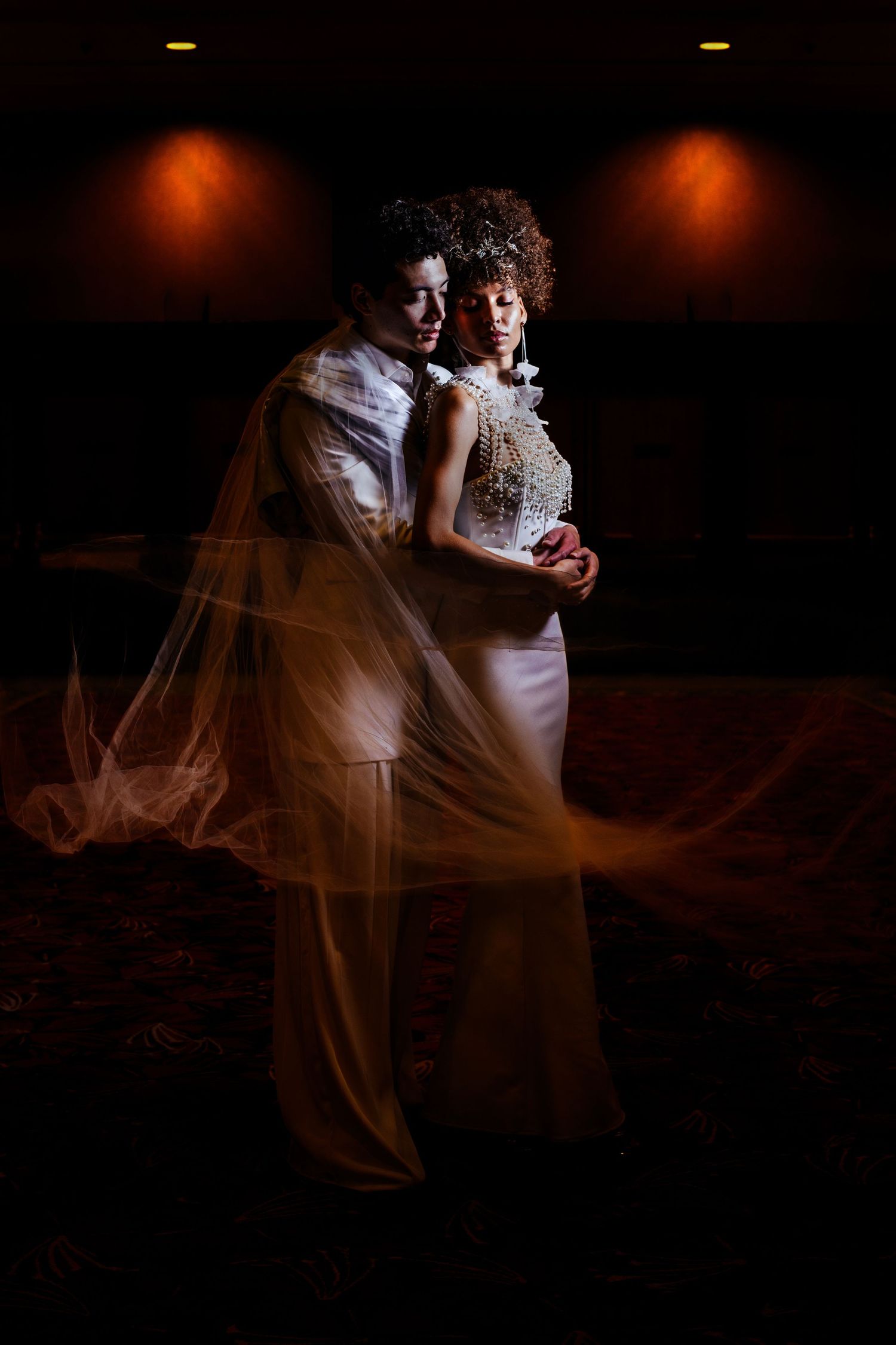Bride and groom share an intimate moment in dramatic lighting, with a flowing veil sweeping elegantly around them.