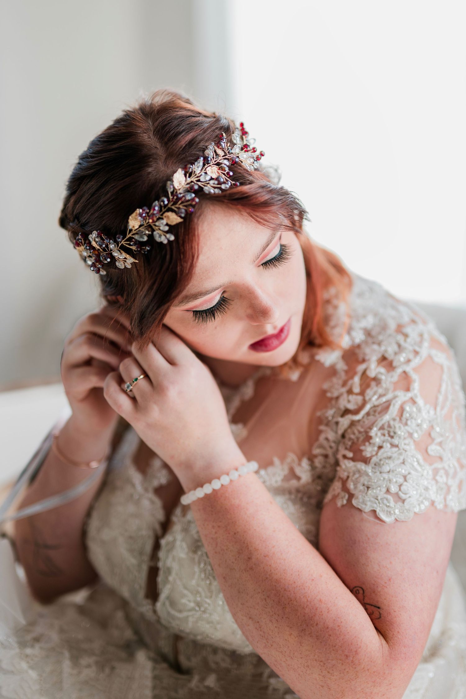 Bride in lace wedding dress wearing floral crown adjusts earring, showcasing elegant bridal hair and makeup.