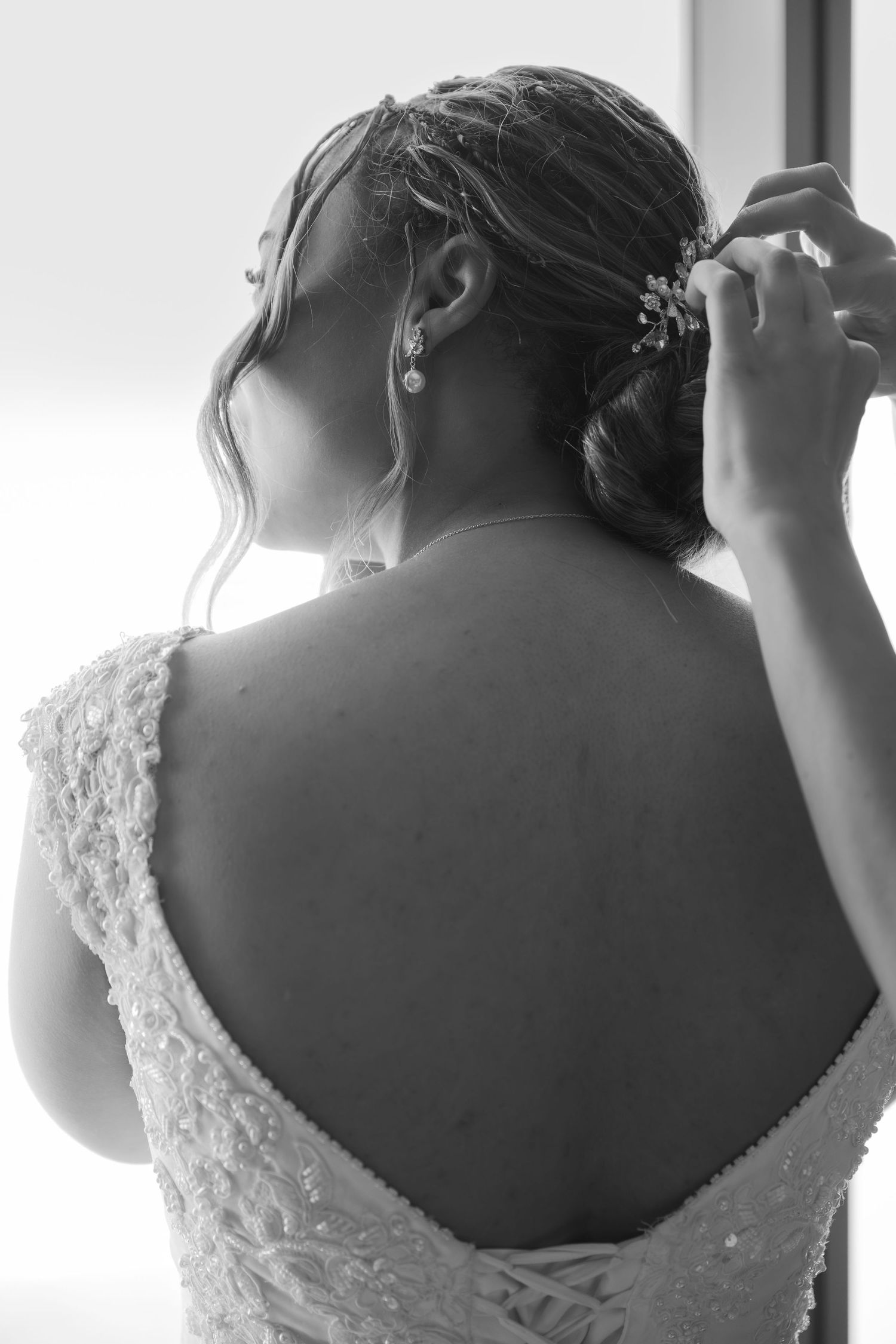 Black and white photo of a bride in a lace gown adjusting her hair accessory, showing her elegant open back.
