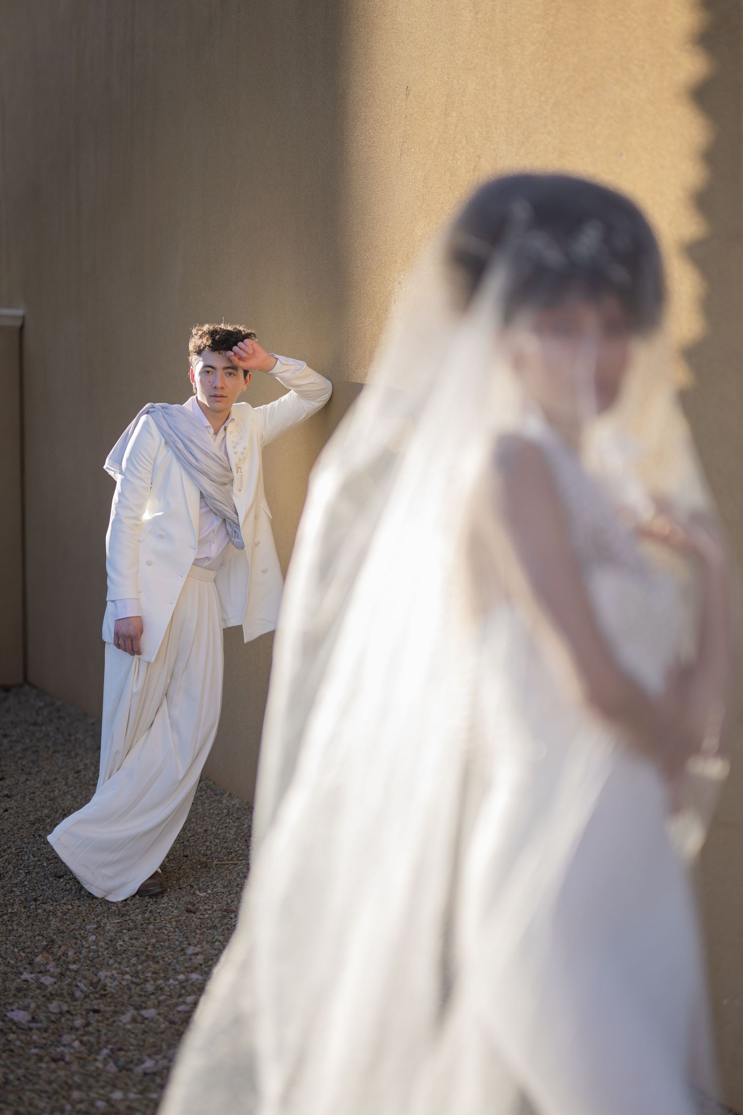 A bride in a white gown and veil stands in the foreground as a groom in a white suit poses against a wall.
