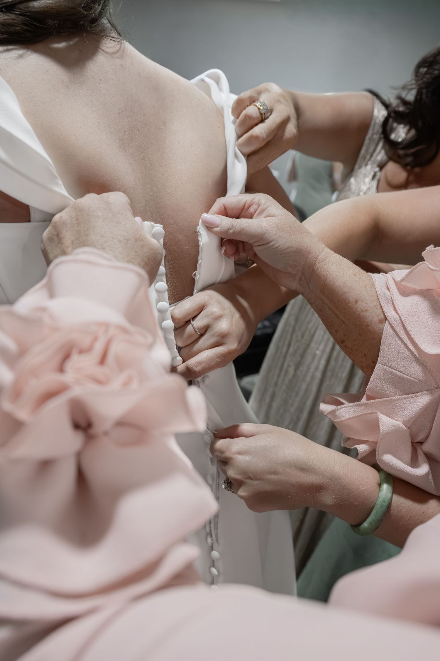 Bridesmaids in pink dresses helping bride button up white wedding gown from behind before ceremony.