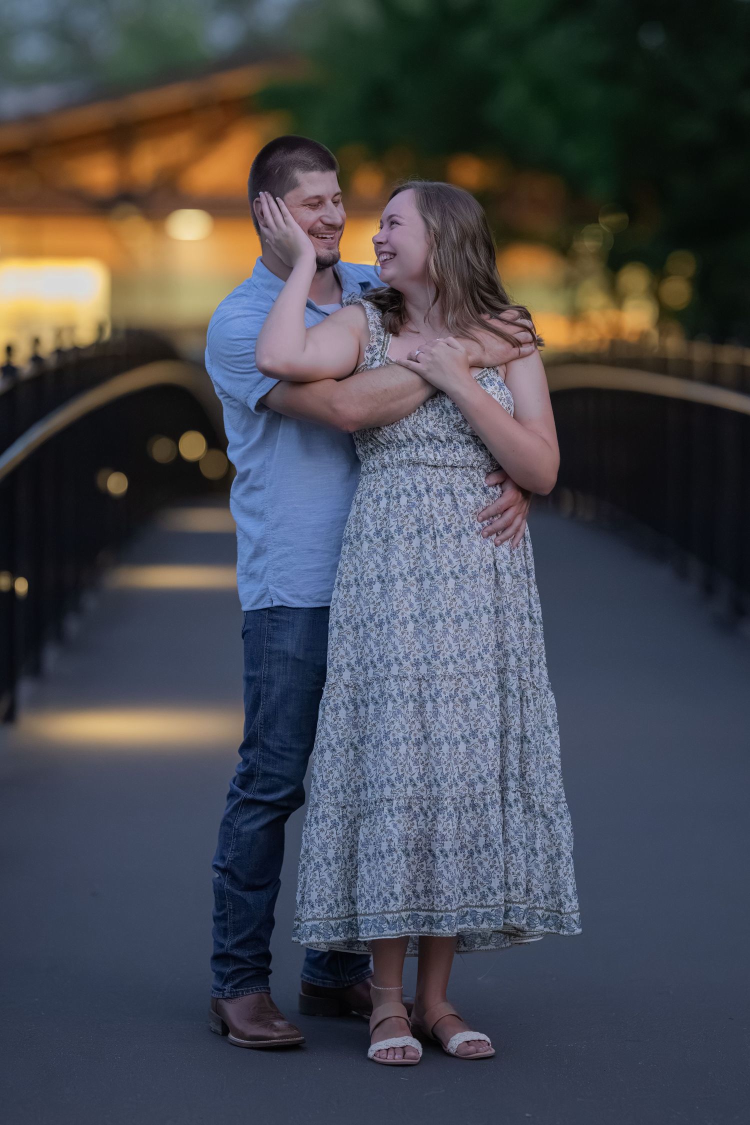 A couple shares a tender moment on a bridge at golden hour, woman in floral dress smiling up at man in blue shirt.