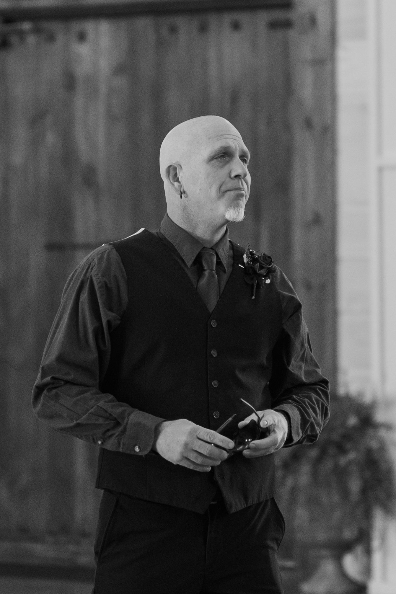 Bald man in black vest and boutonniere holds sunglasses outdoors near wooden barn doors in black and white photo.