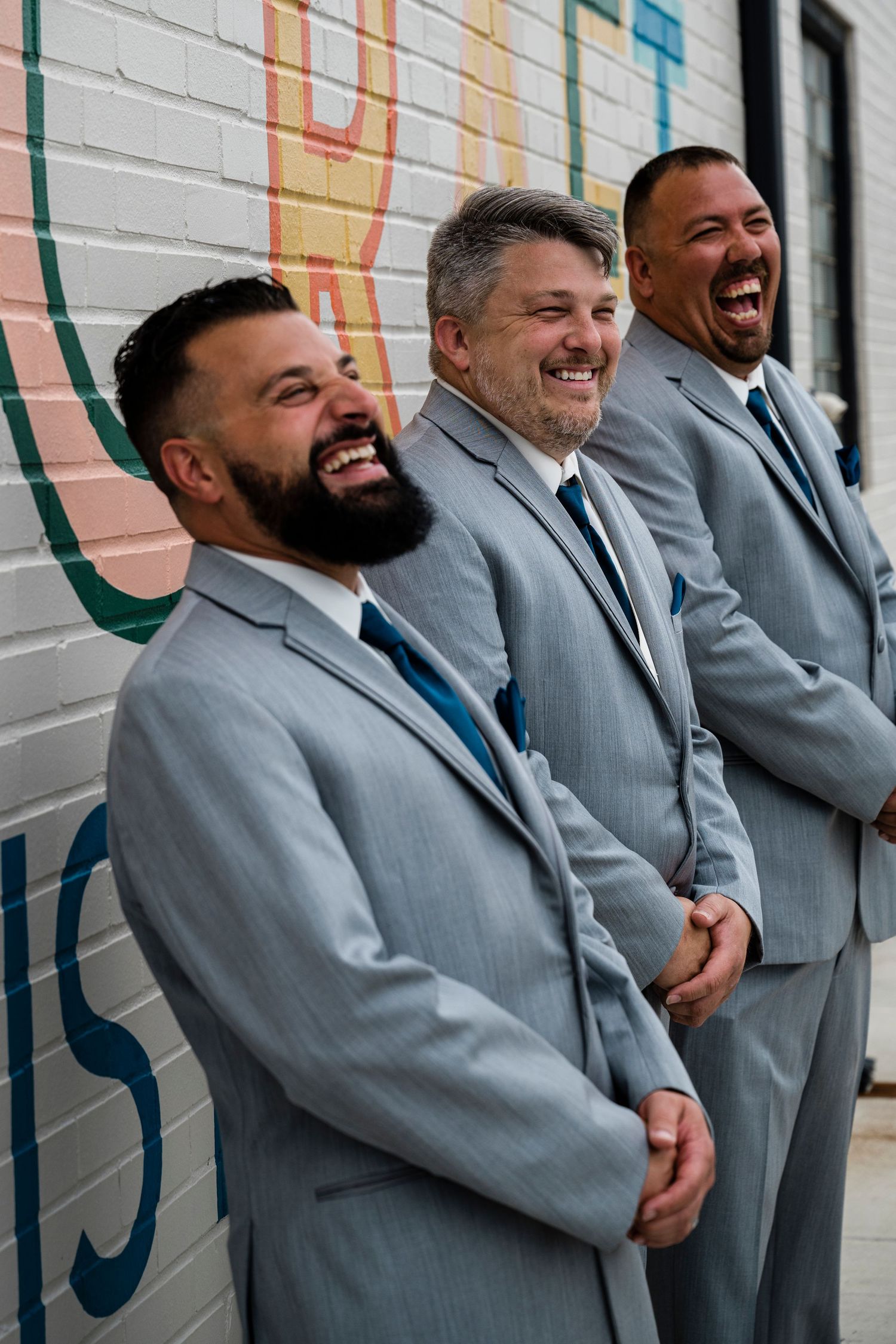 Three men in matching gray suits laughing joyfully together against a colorful mural wall.