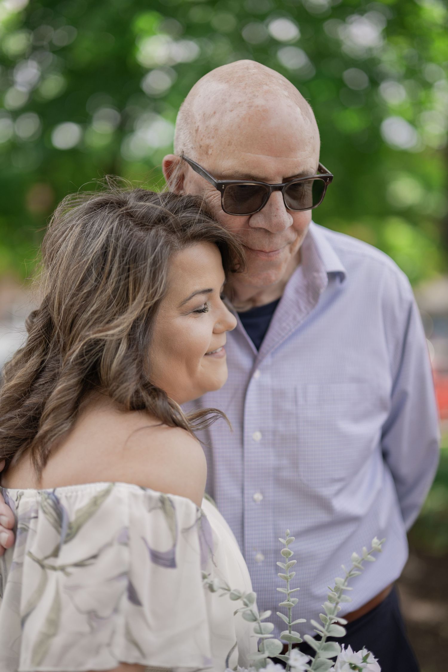 Older man in sunglasses tenderly embraces a woman in an off-shoulder floral dress outdoors.